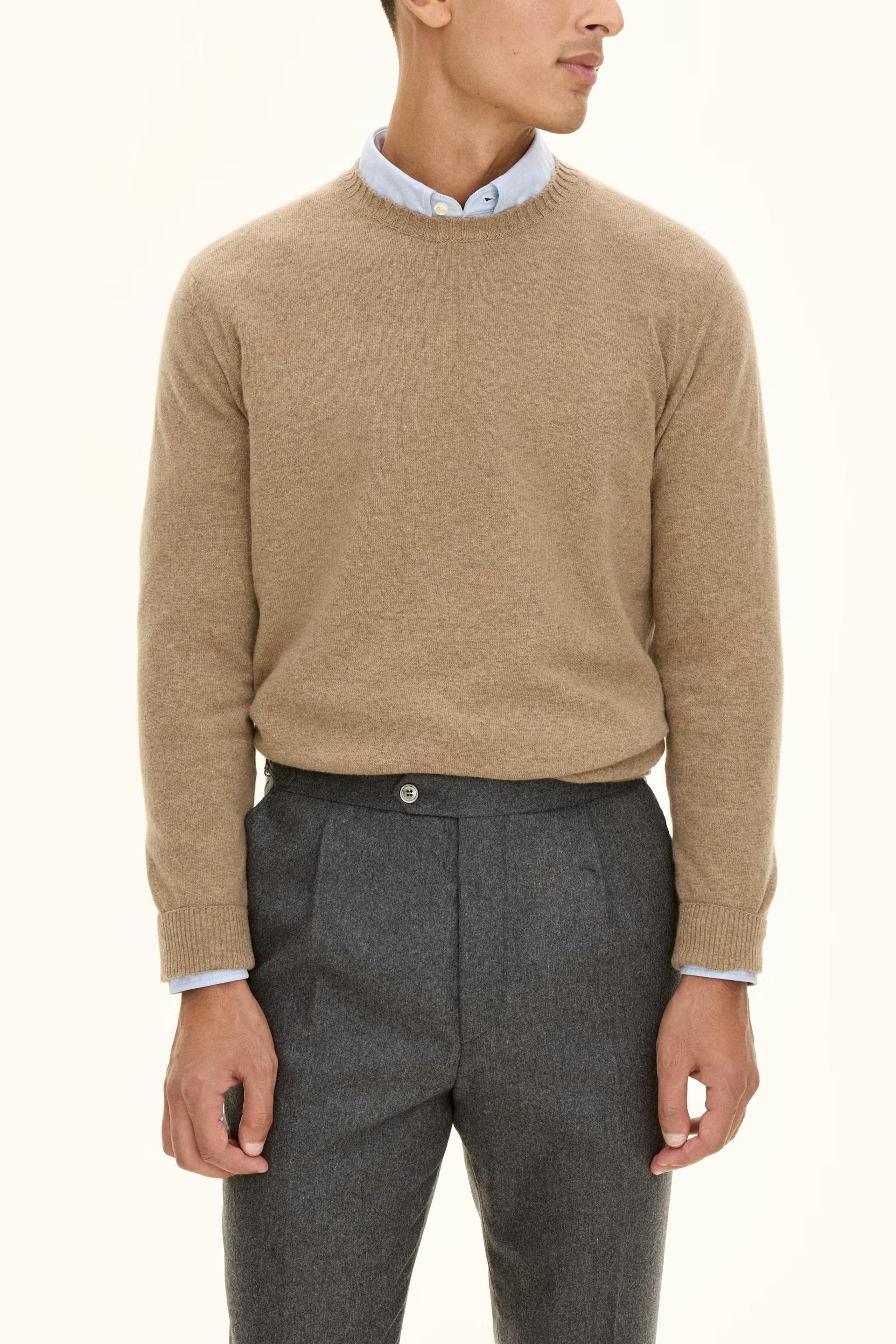 A man wears a camel crew-neck sweater over a light blue shirt, paired with grey pleated trousers.