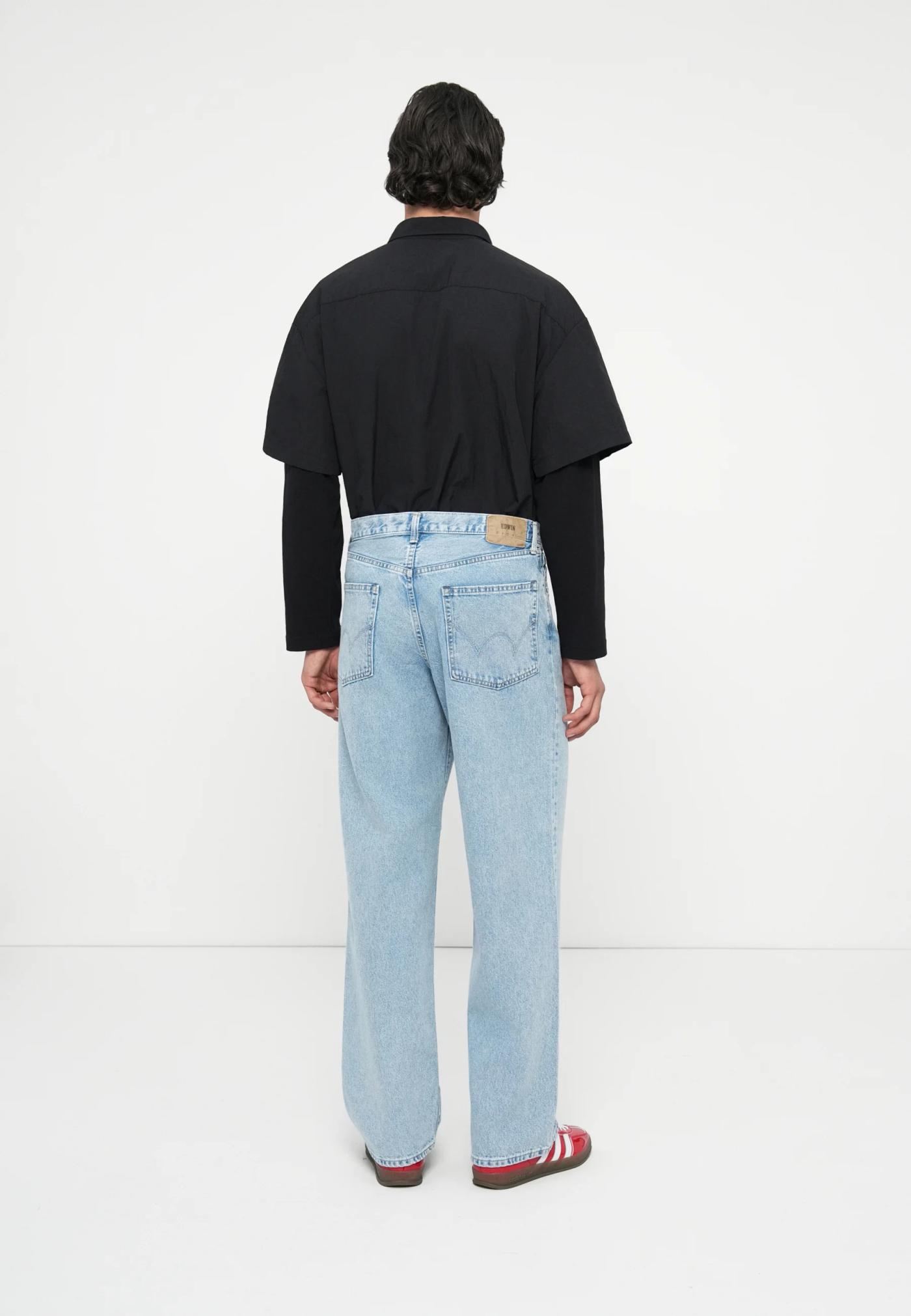 A person seen from behind, wearing a black layered shirt, light wash baggy jeans, and red sneakers.