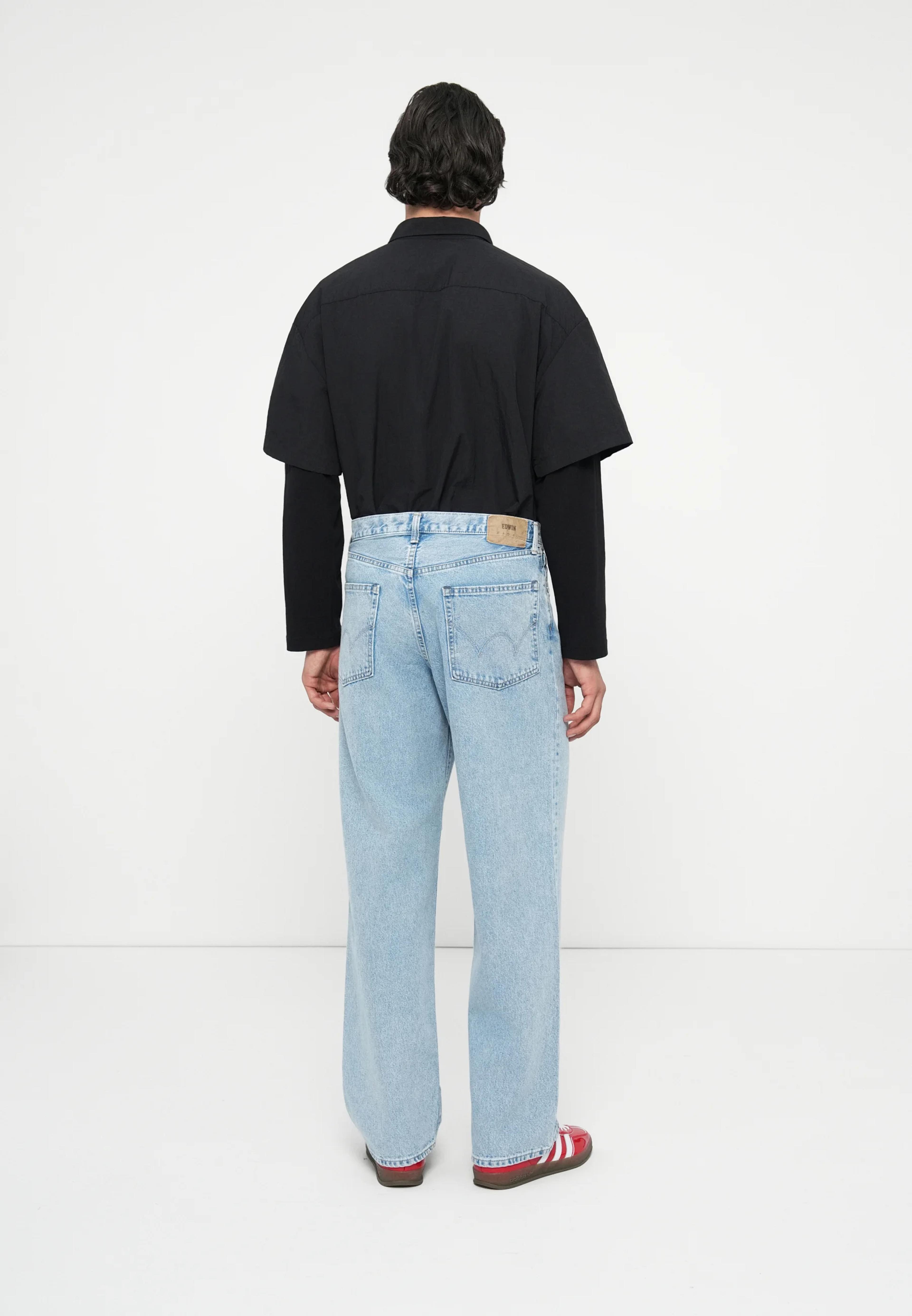 A person seen from behind, wearing a black layered shirt, light wash baggy jeans, and red sneakers.