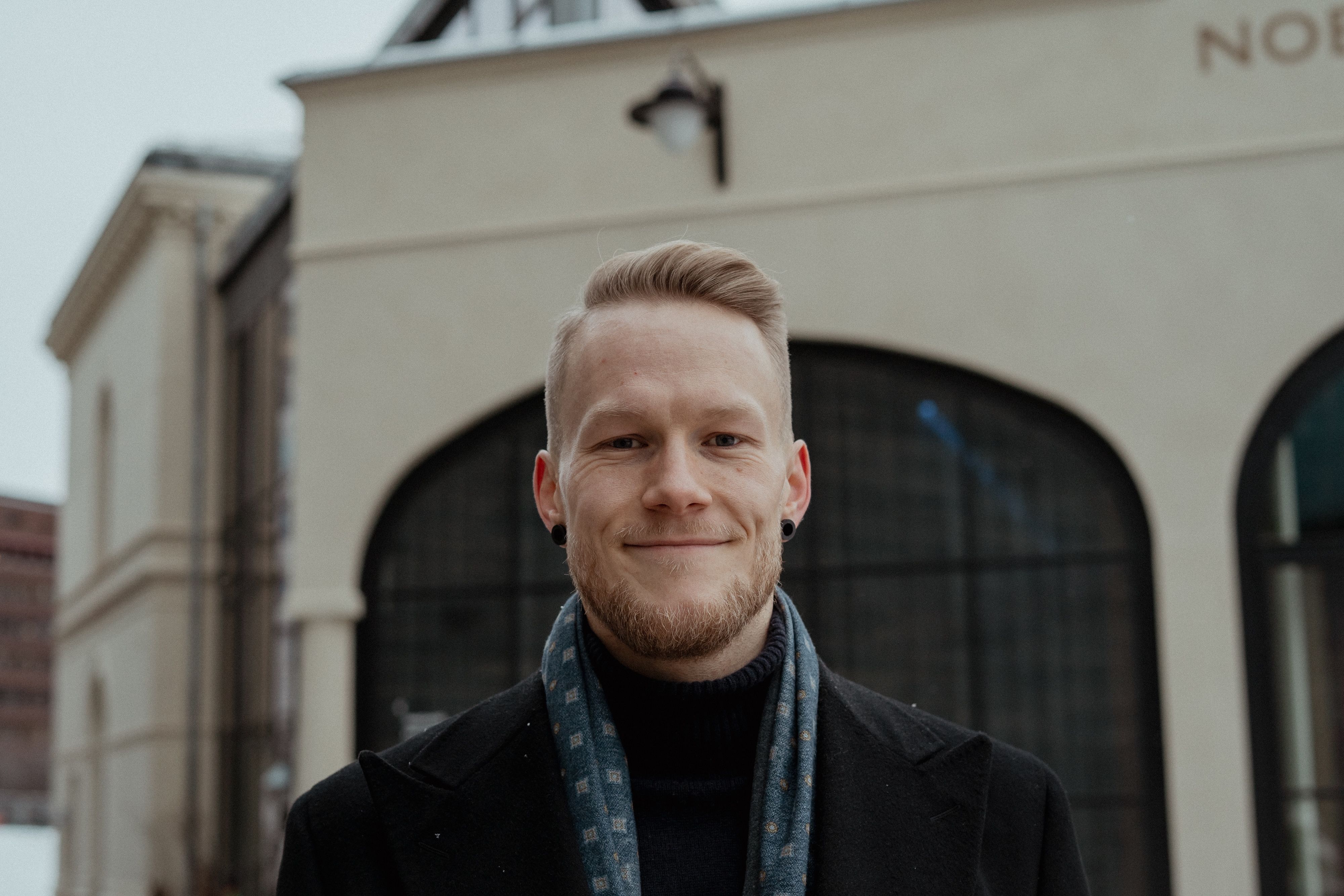 A smiling man with light hair and a beard, wearing a black coat and blue scarf, stands in front of a light-colored building.