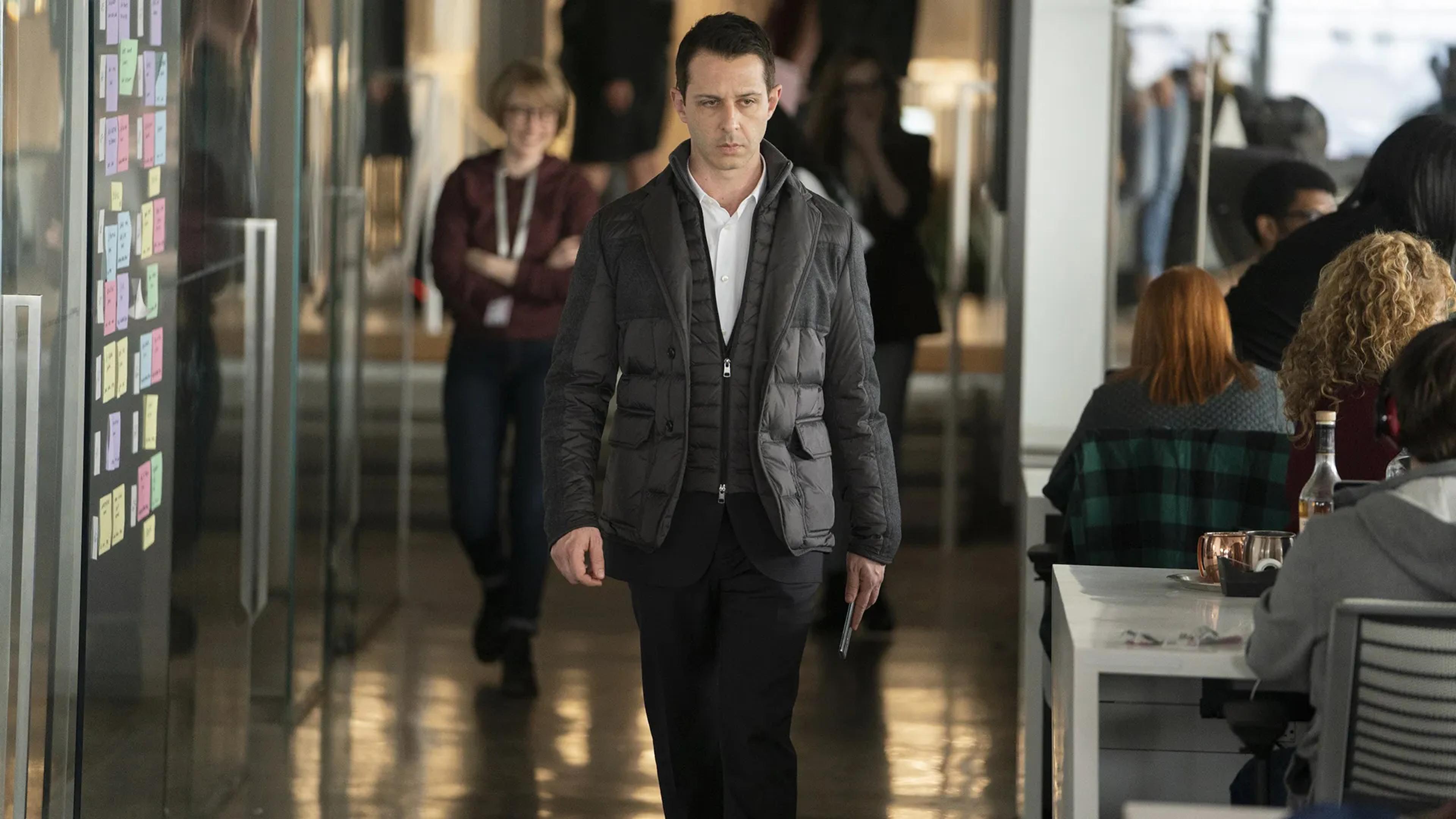 A man in a dark puffer jacket walks through a bright modern office with people seated at tables.