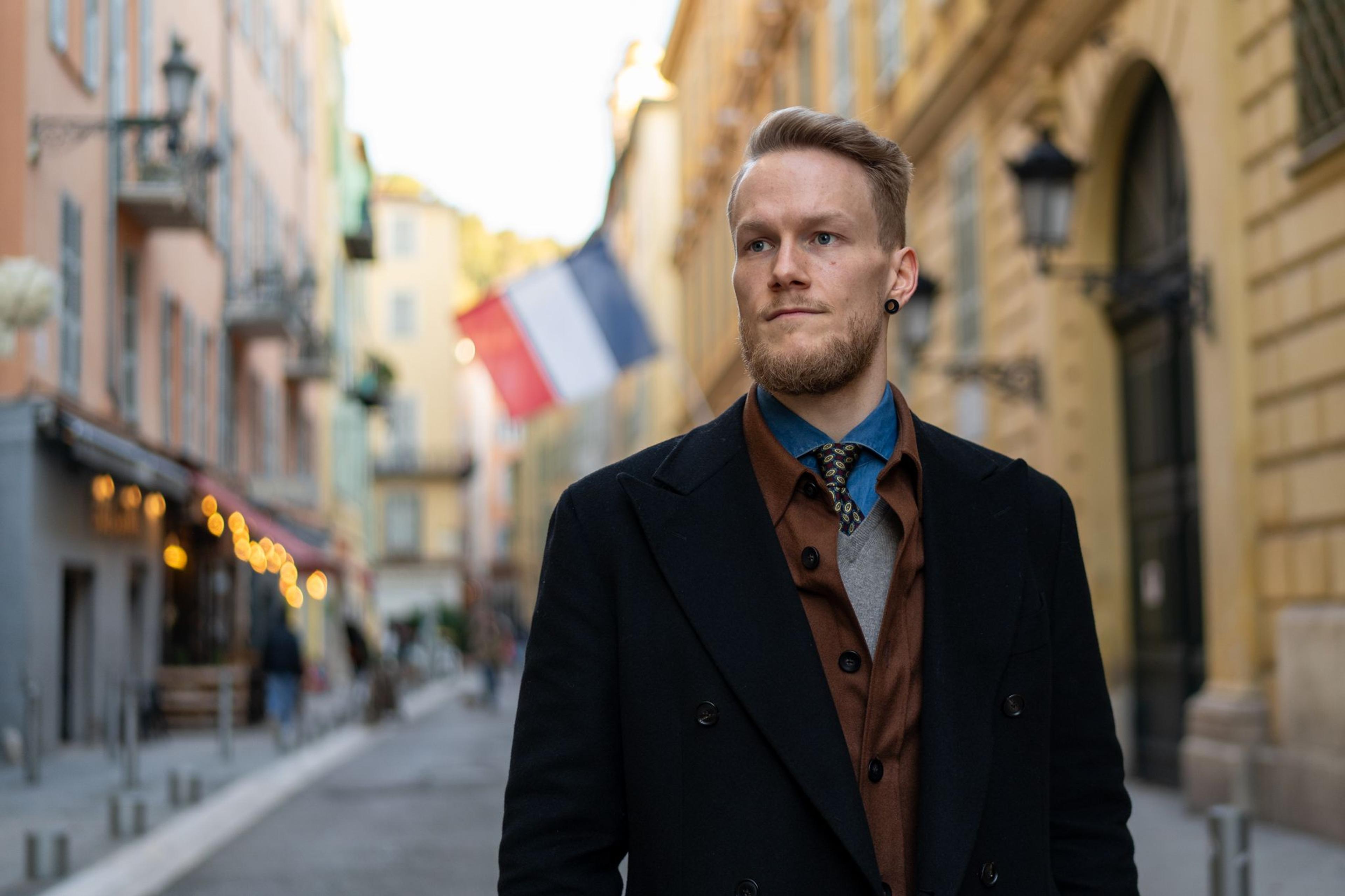 A blonde man with a beard wearing a black coat stands on a colorful street with a French flag in the background.