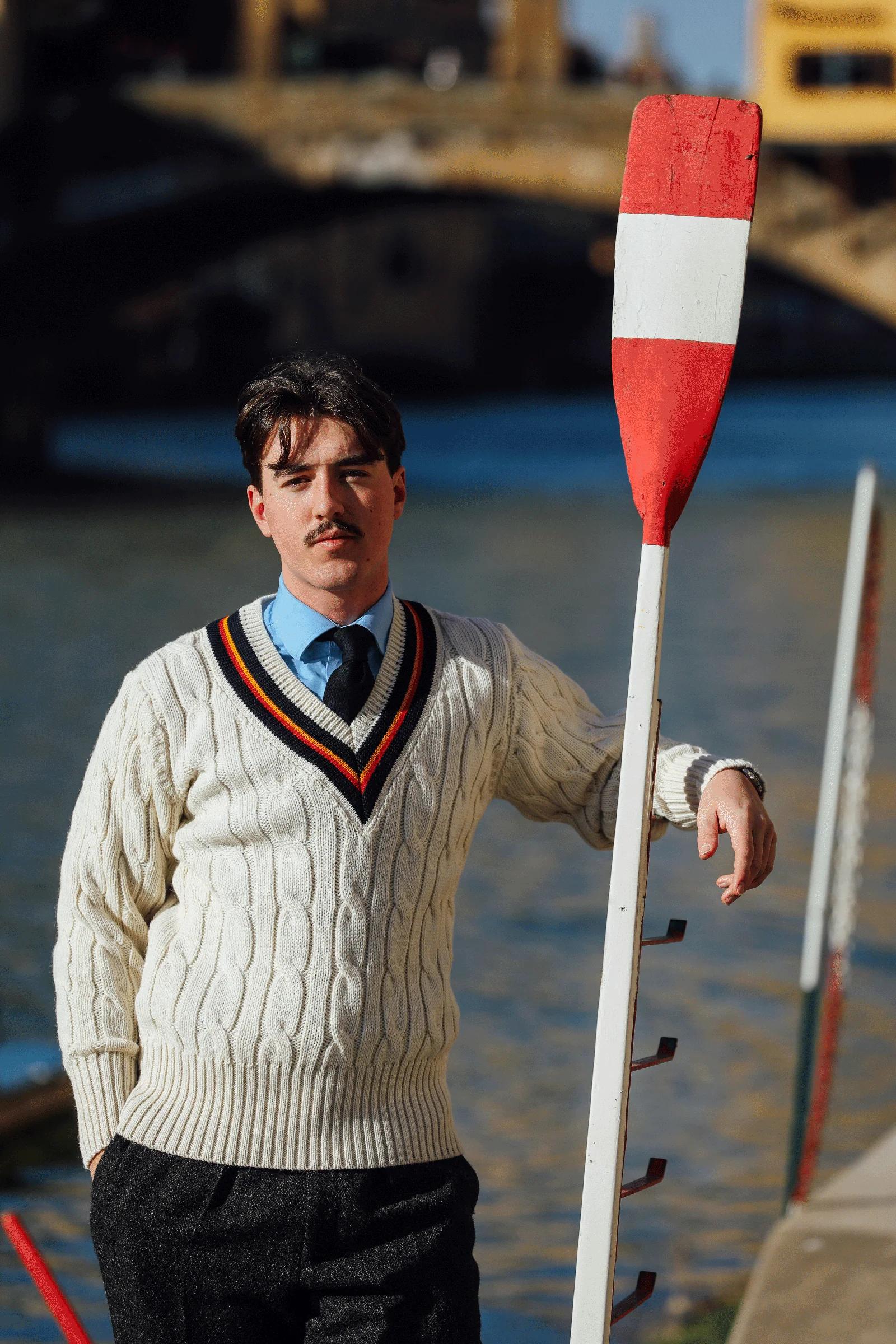 A man with a mustache in a cream cable-knit sweater leans on a red and white oar by a river.