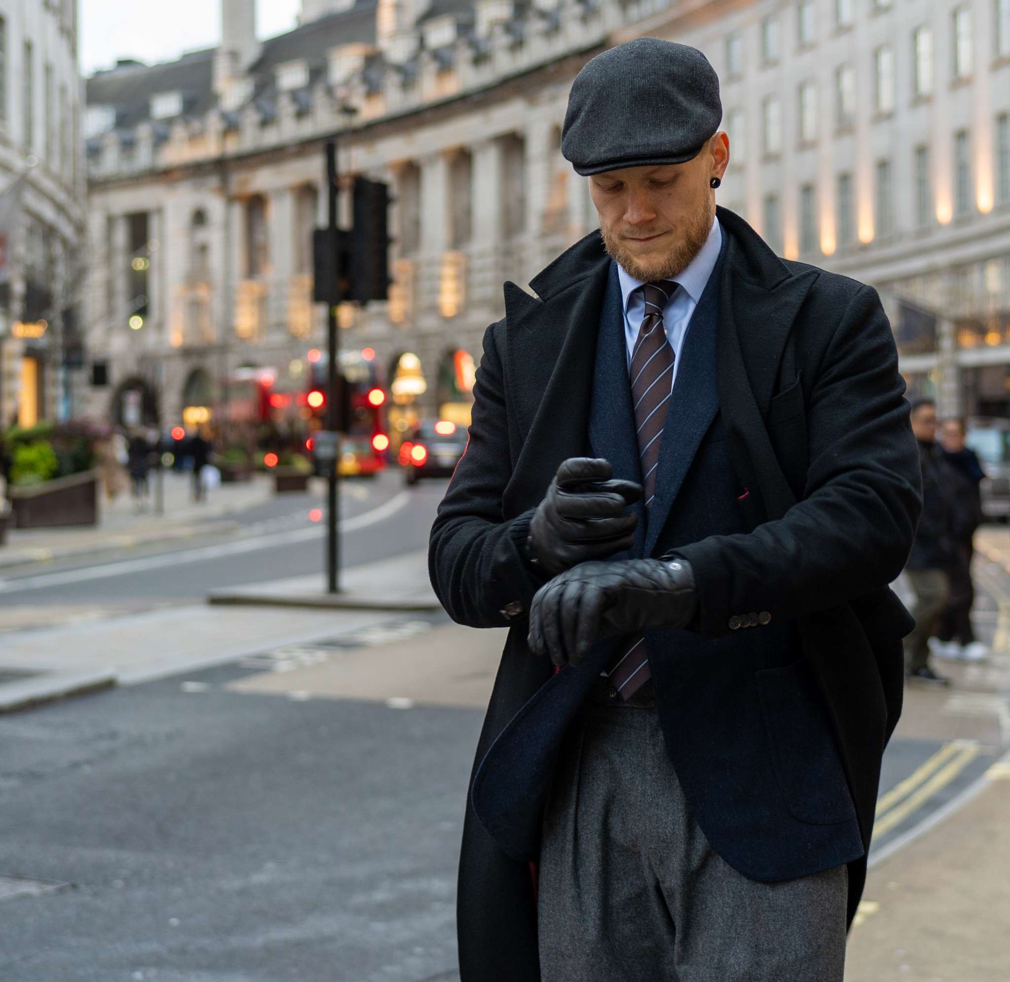 Stylish man in a flat cap, black coat, and gloves adjusting his wrist on a city street.
