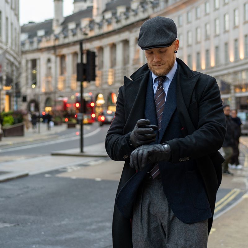 Stylish man in a flat cap, black coat, and gloves adjusting his wrist on a city street.
