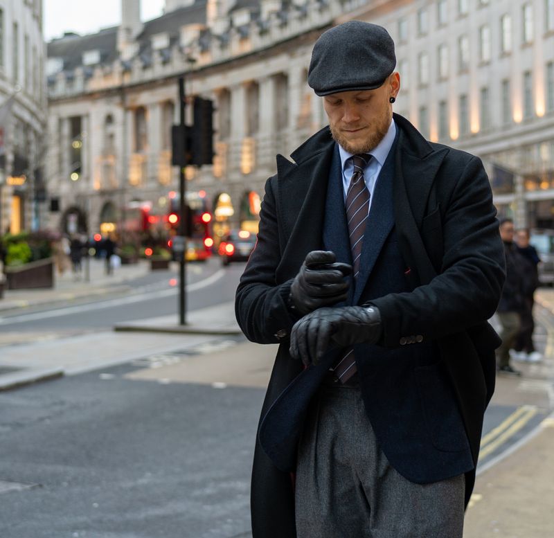 Stylish man in a flat cap, black coat, and gloves adjusting his wrist on a city street.