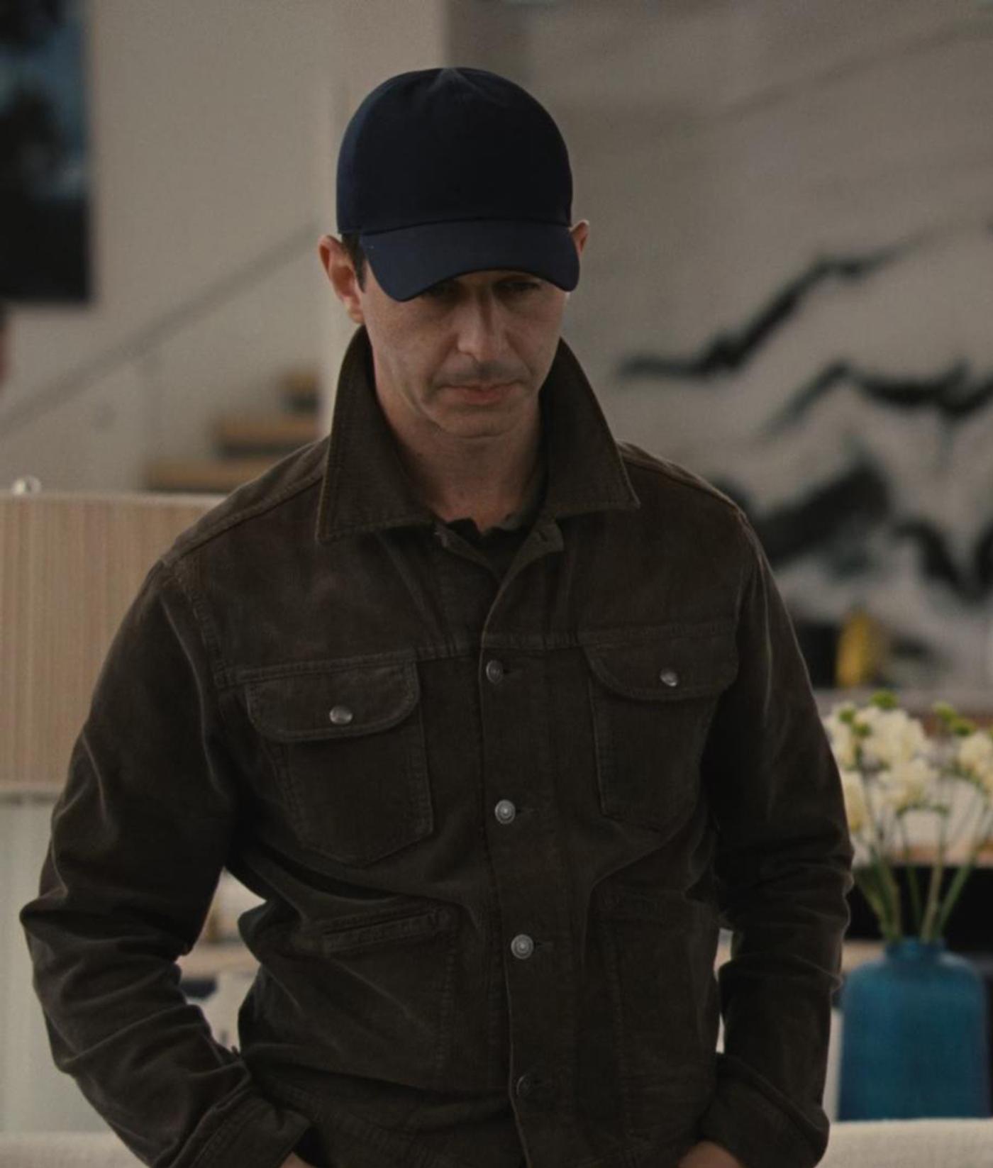 A man in a dark blue baseball cap and dark brown corduroy jacket looks down with a somber expression.