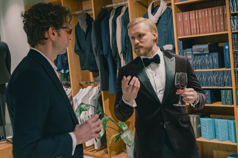 Two men, one in a tuxedo holding wine and the other in a suit, conversing in an upscale clothing store.