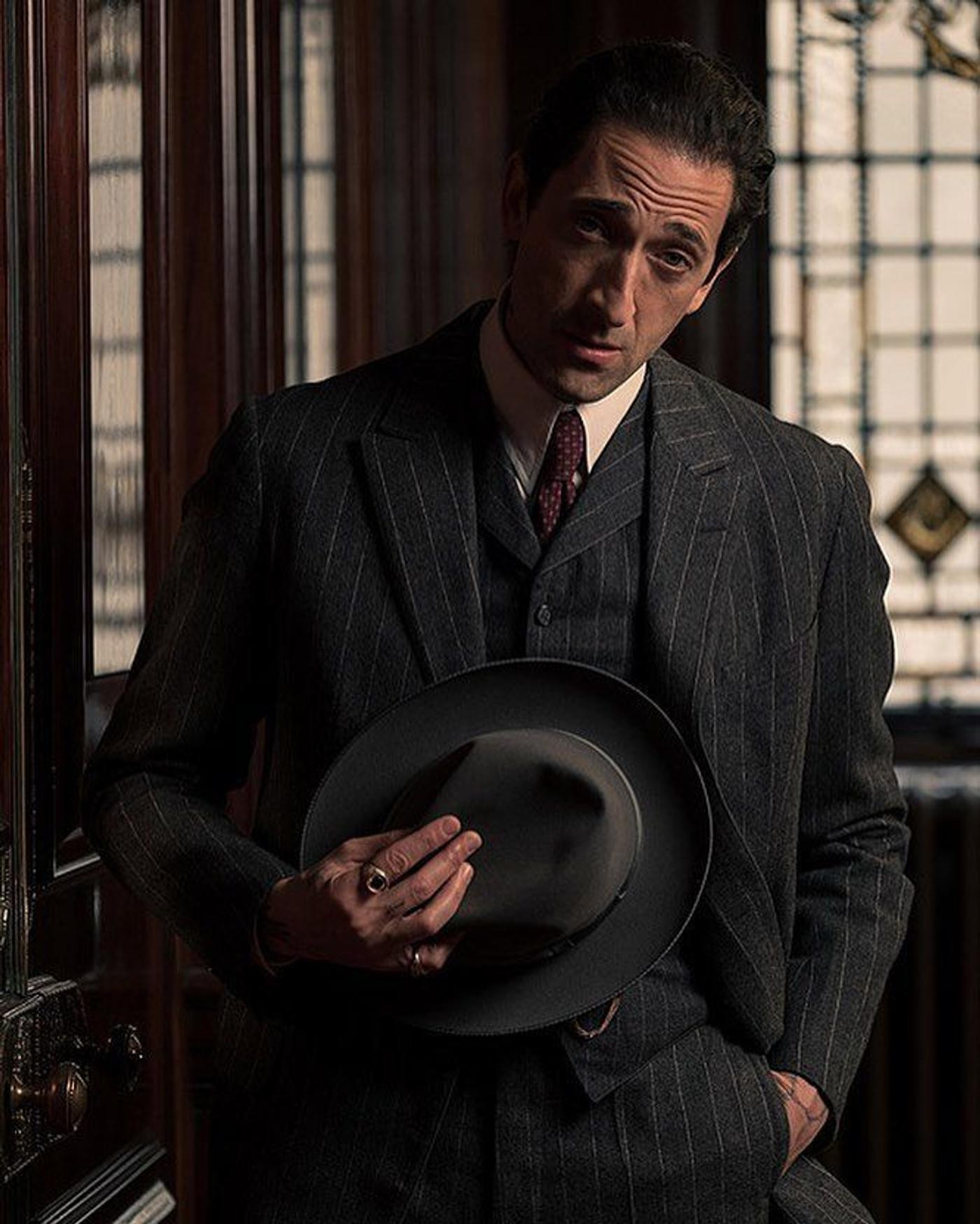 Adrien Brody in a dark pinstripe suit holds a fedora.