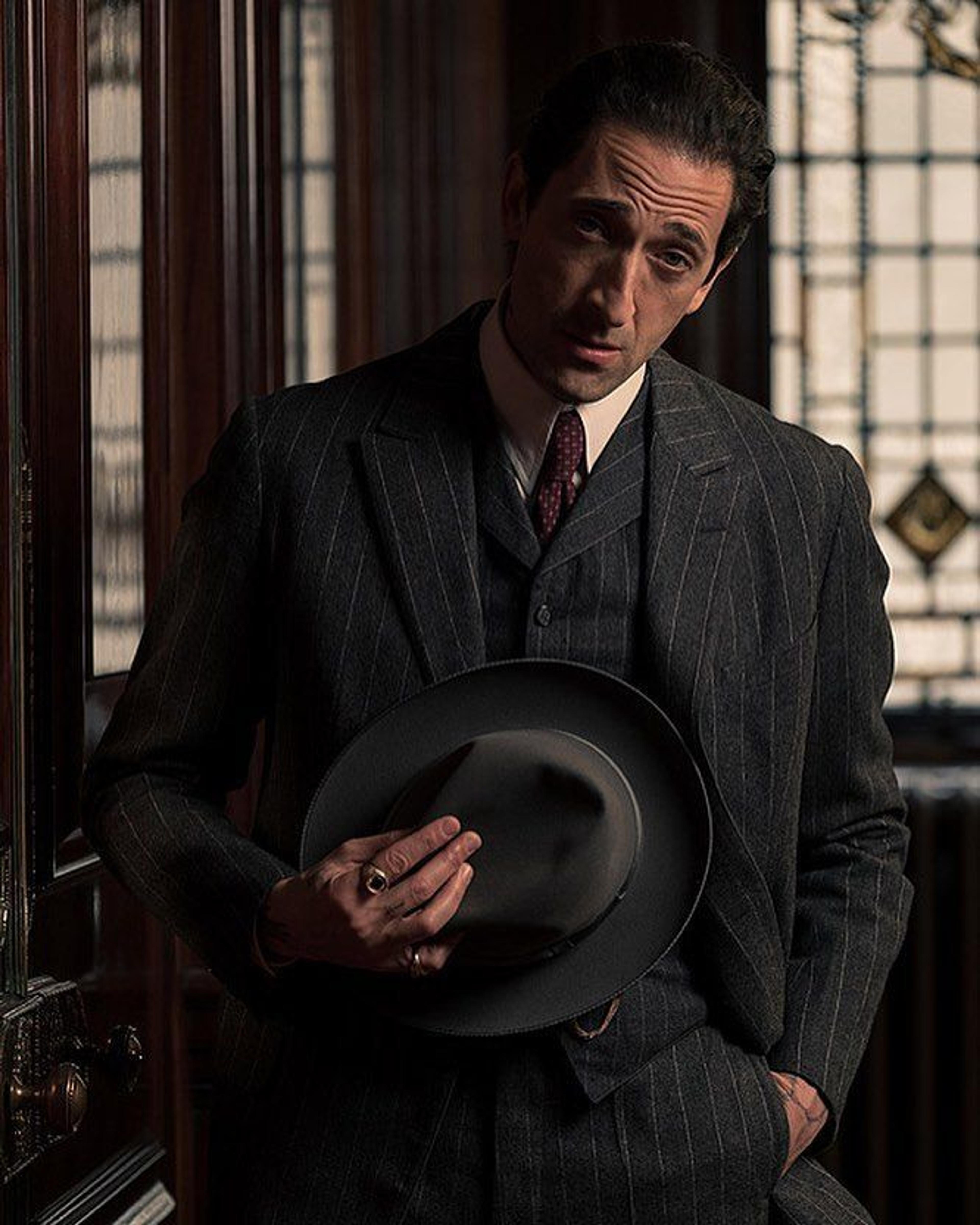 Adrien Brody in a dark pinstripe suit holds a fedora.