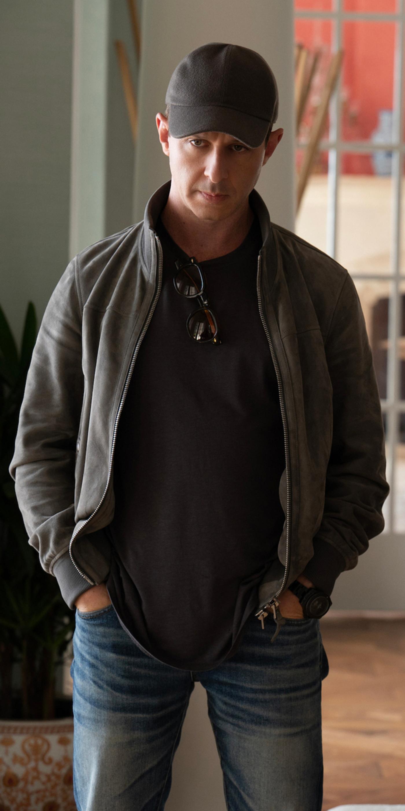 A man in a dark baseball cap, open gray jacket with sunglasses, and jeans, hands in pockets.