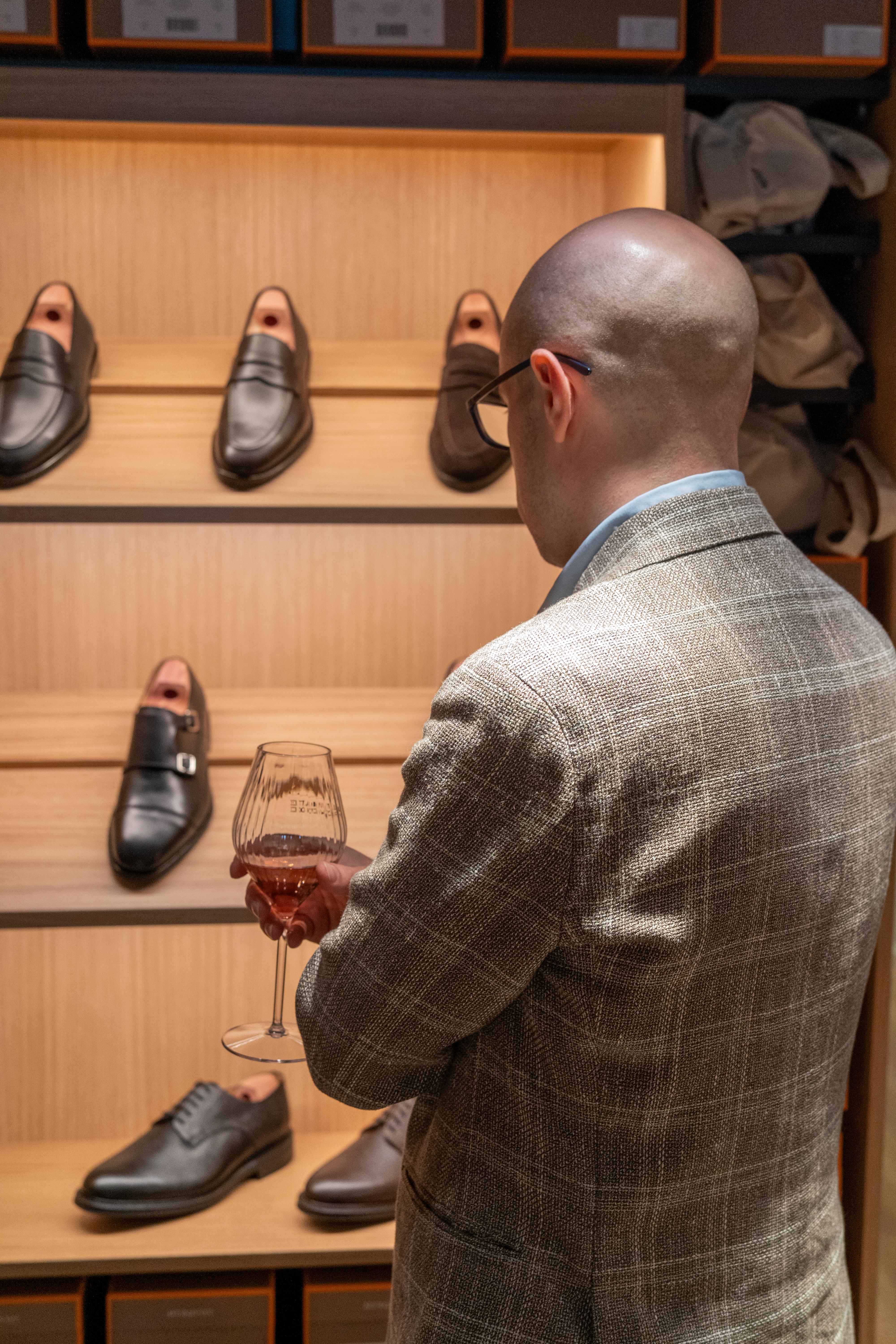 a man in a suit holds a glass of wine in front of a display of shoes