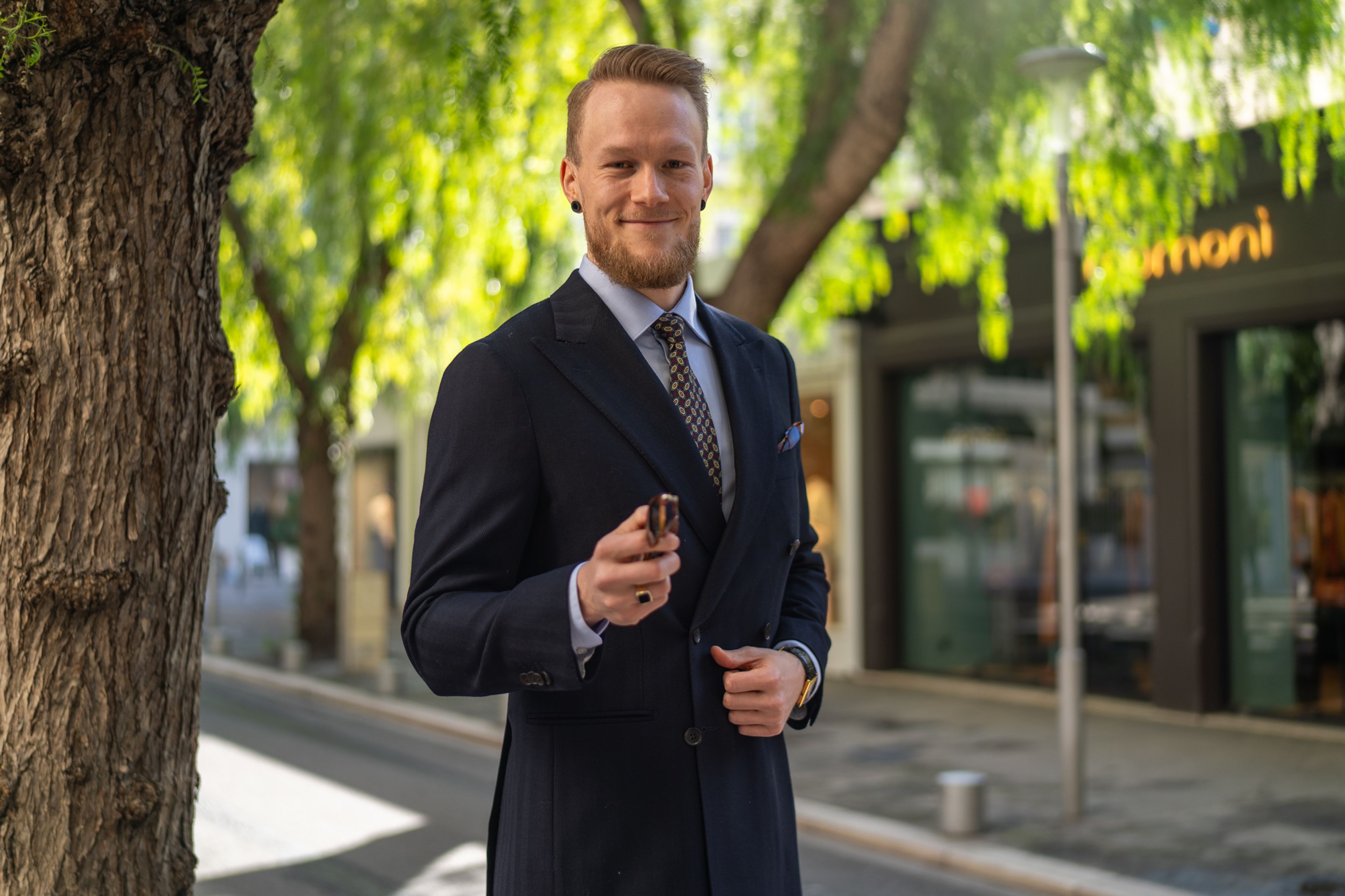 A smiling man in a navy double-breasted suit holds sunglasses on a sunny street.