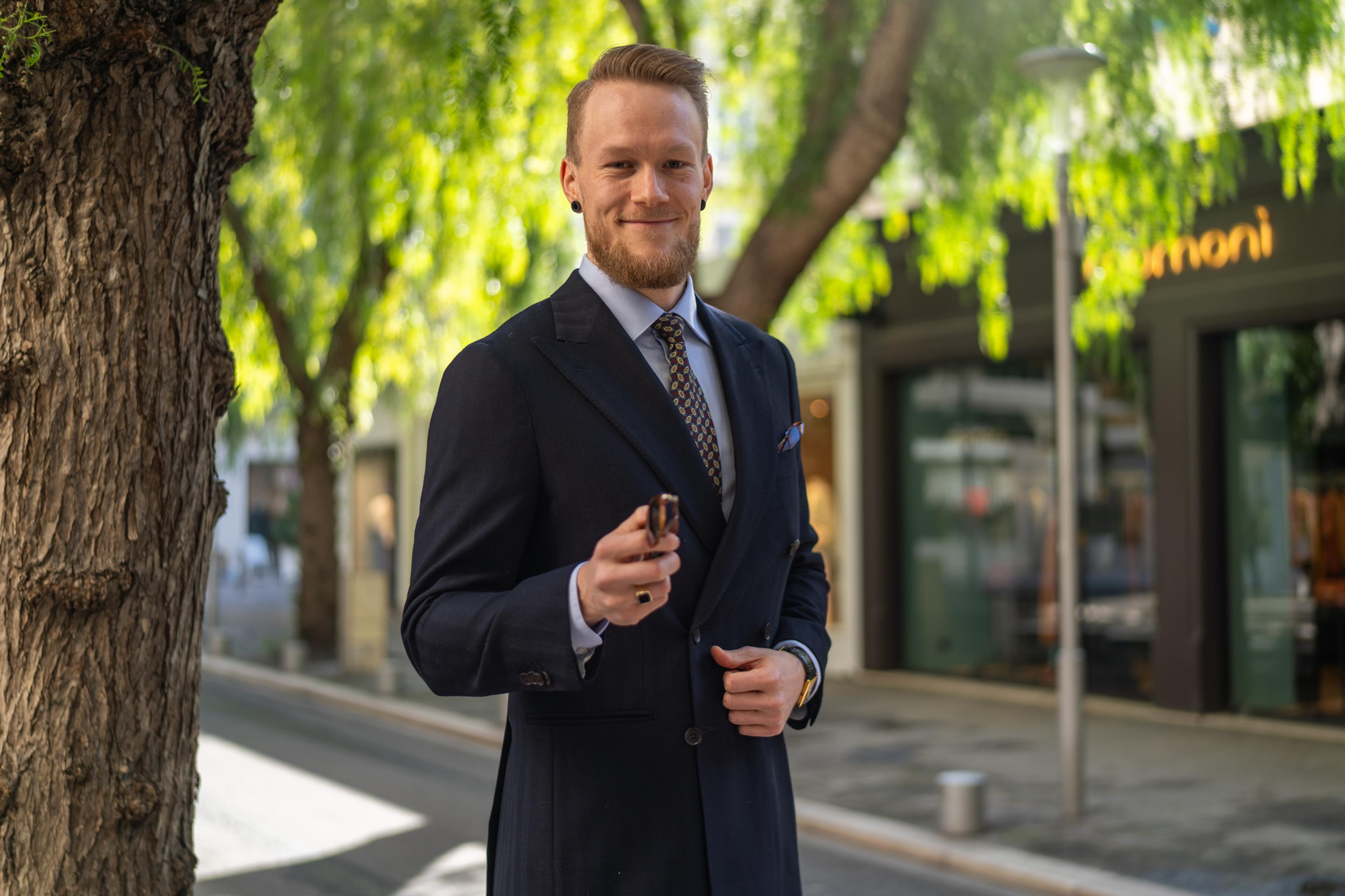 A smiling man in a navy double-breasted suit holds sunglasses on a sunny street.