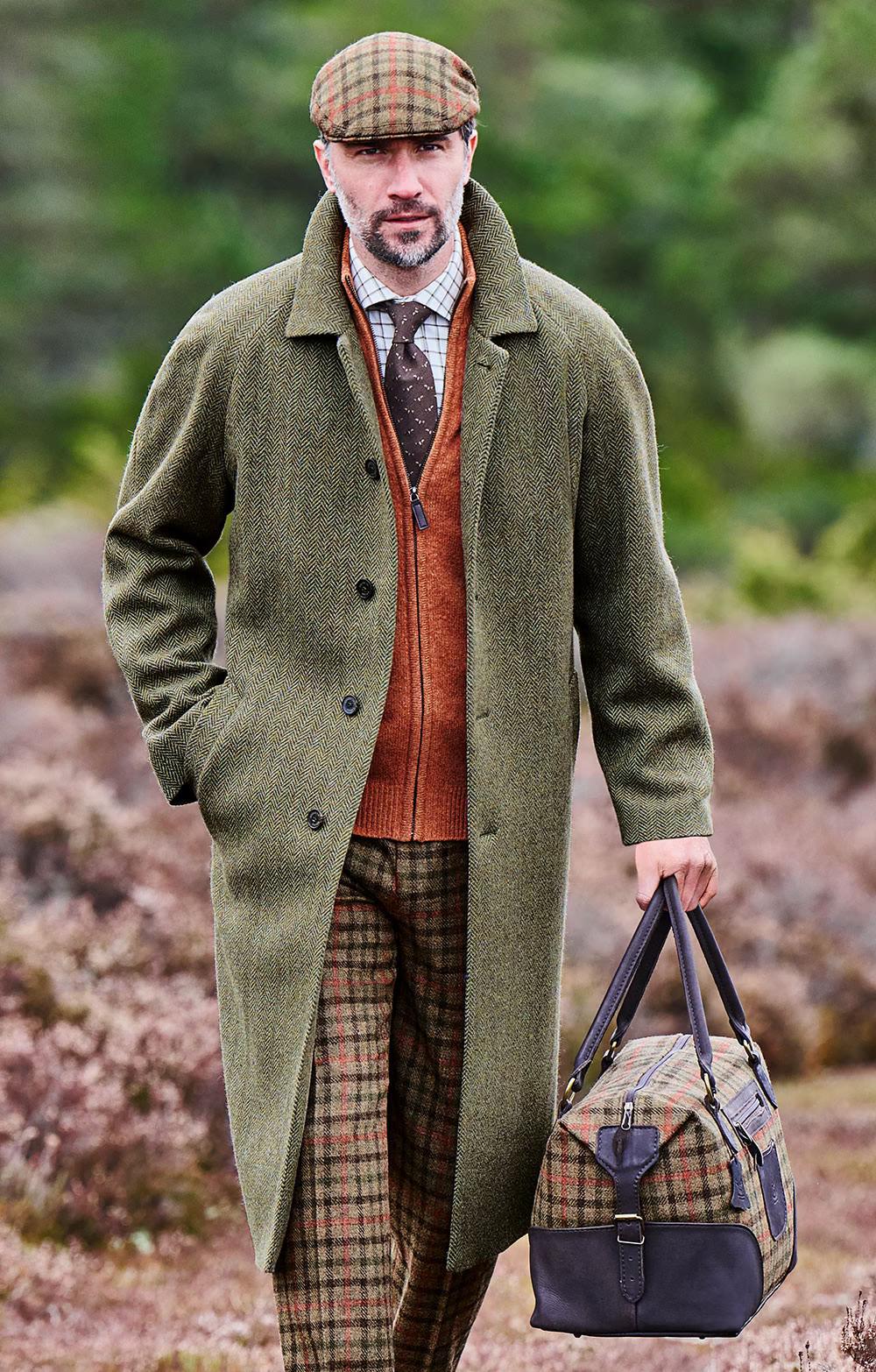 Man in a green tweed overcoat, plaid cap, and trousers, carrying a matching plaid travel bag, walking outdoors.
