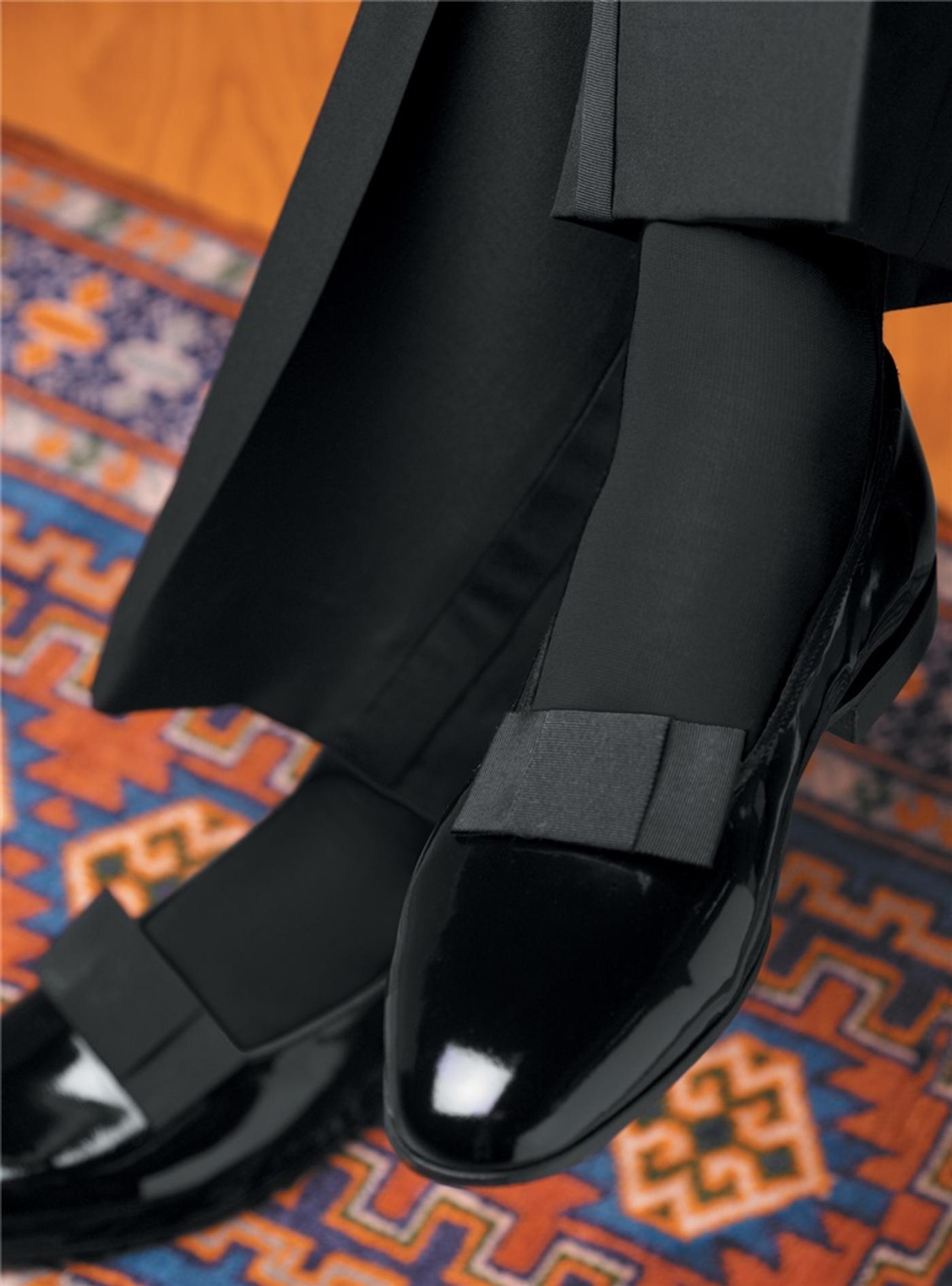 Black tuxedo pants and shiny patent leather dress shoes with a grosgrain bow on a patterned rug.