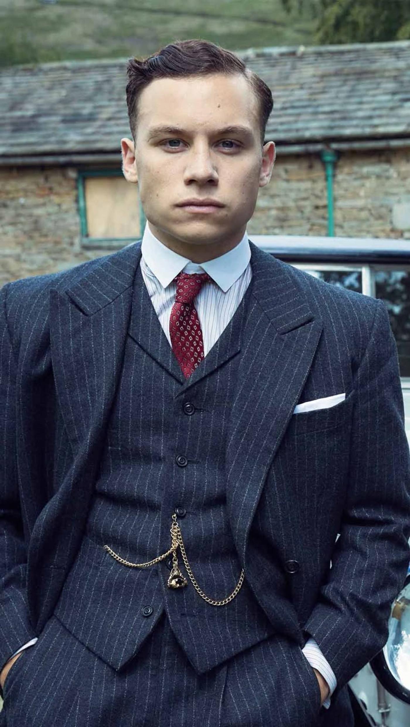 A young man in a dark pinstripe three-piece suit with a red tie and pocket watch chain looks seriously at the camera.