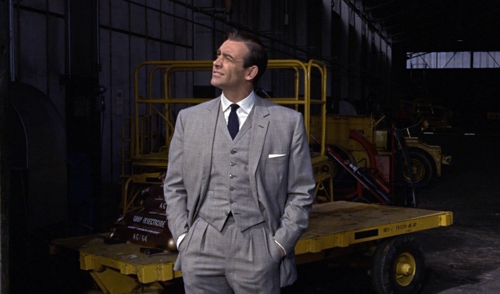 Sean Connery as James Bond in a light grey suit, looking up in an industrial hangar.