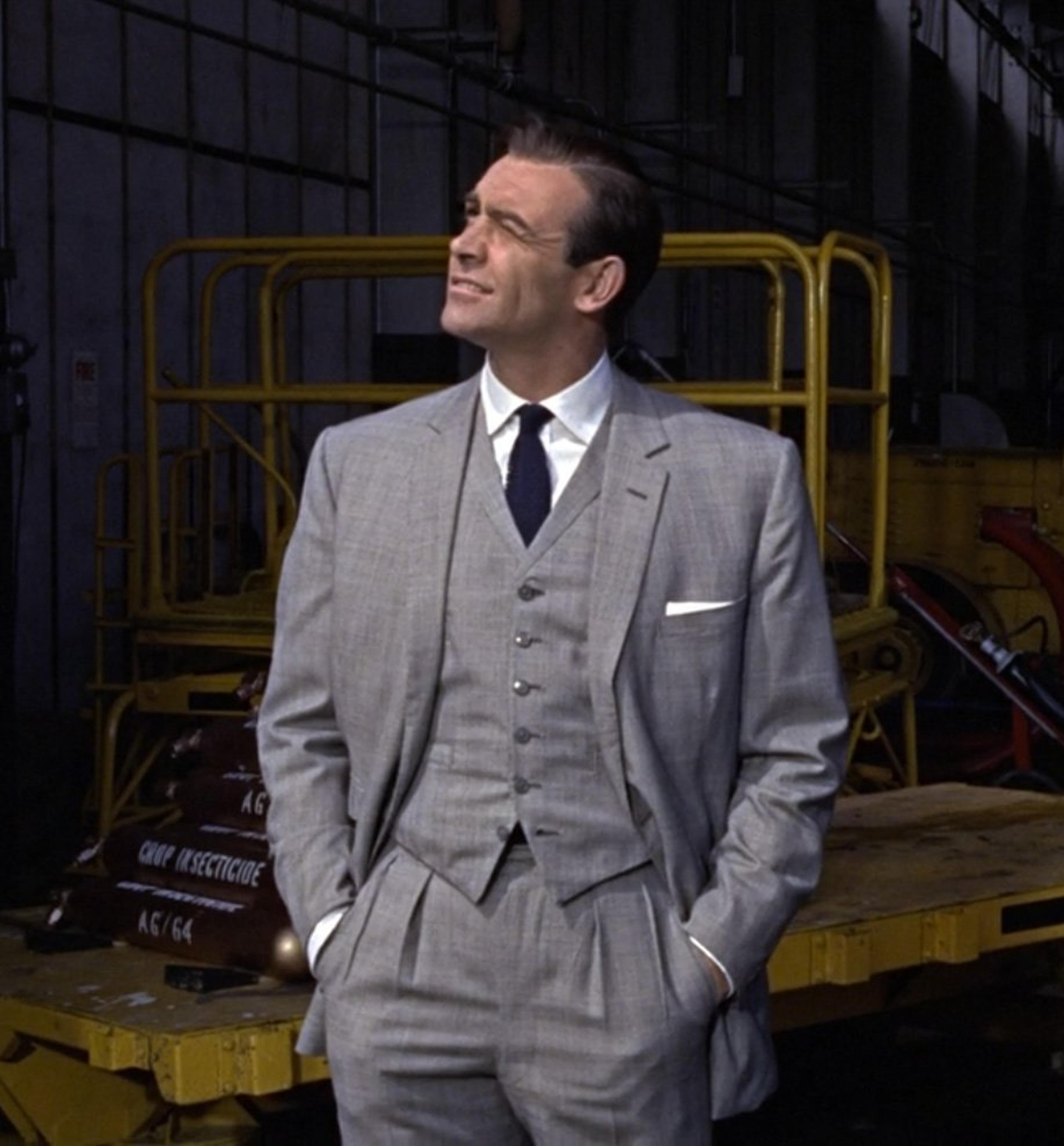Sean Connery as James Bond in a light grey suit, looking up in an industrial hangar.