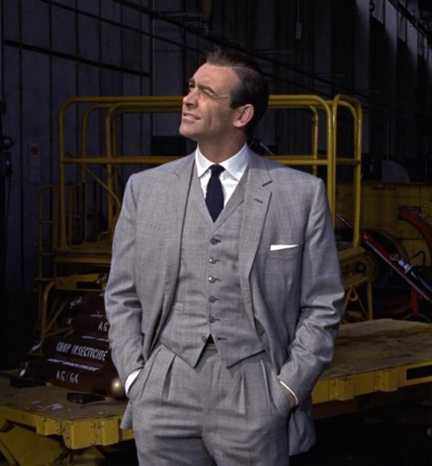 Sean Connery as James Bond in a light grey suit, looking up in an industrial hangar.