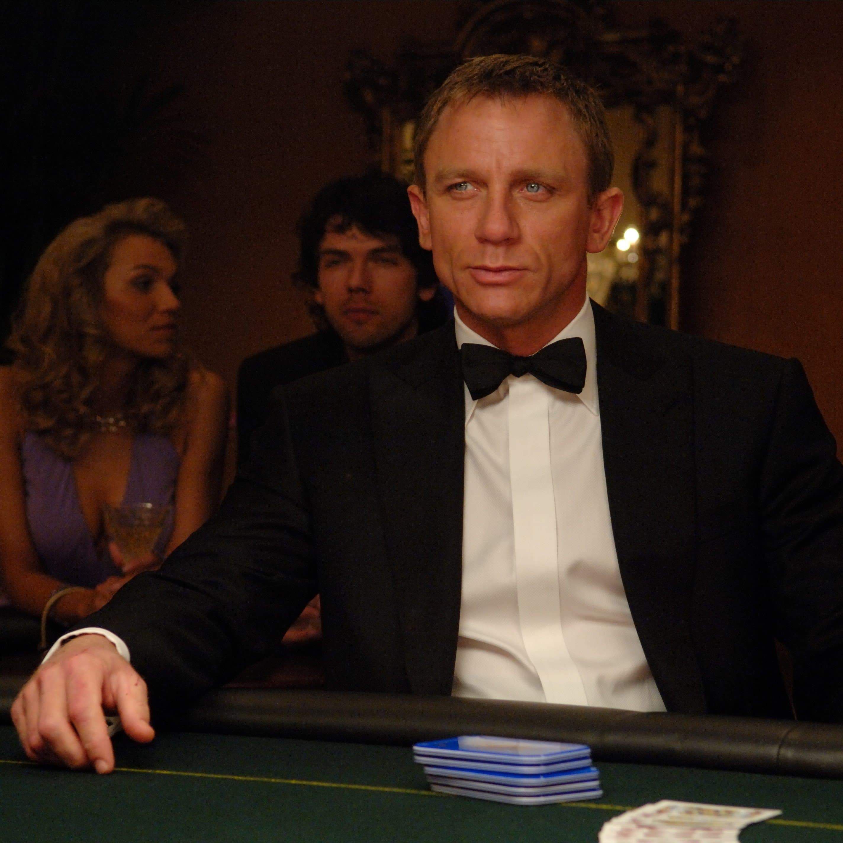 Daniel Craig in a black tuxedo sitting at a poker table, looking intently, with other people in the background.