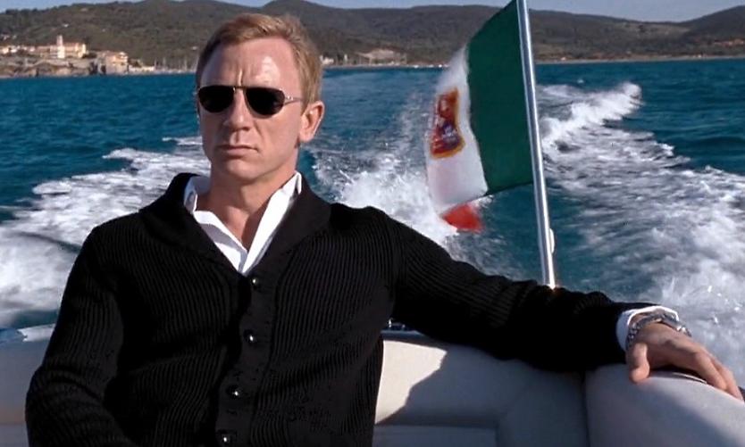 Daniel Craig in sunglasses and a black cardigan on a boat with an Italian flag.