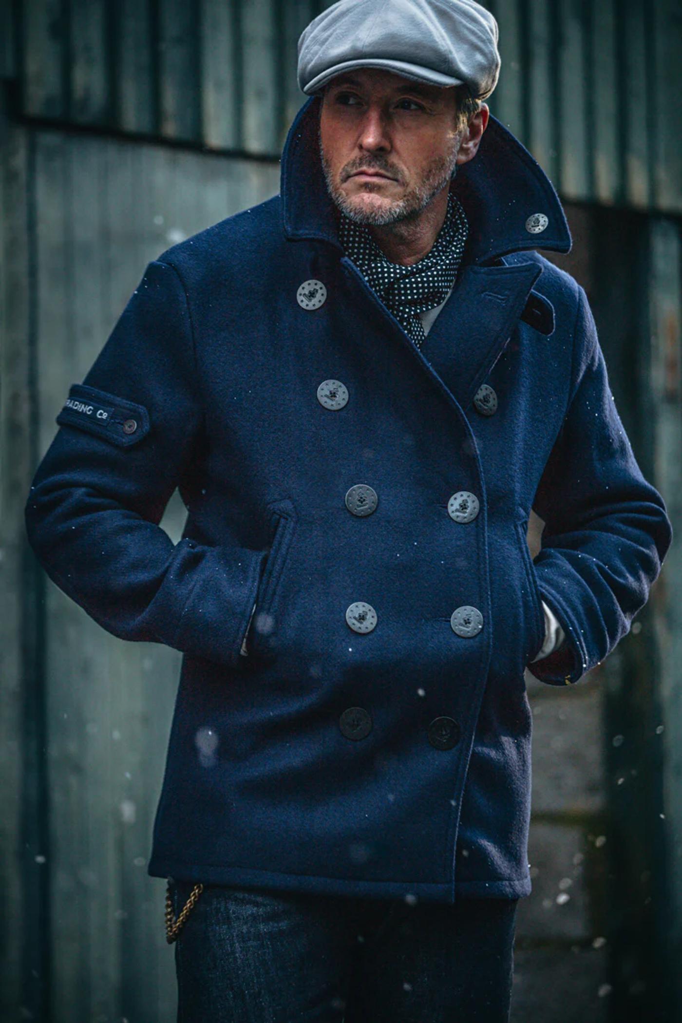A man in a dark blue pea coat, grey cap, and polka dot scarf, with snowflakes falling.