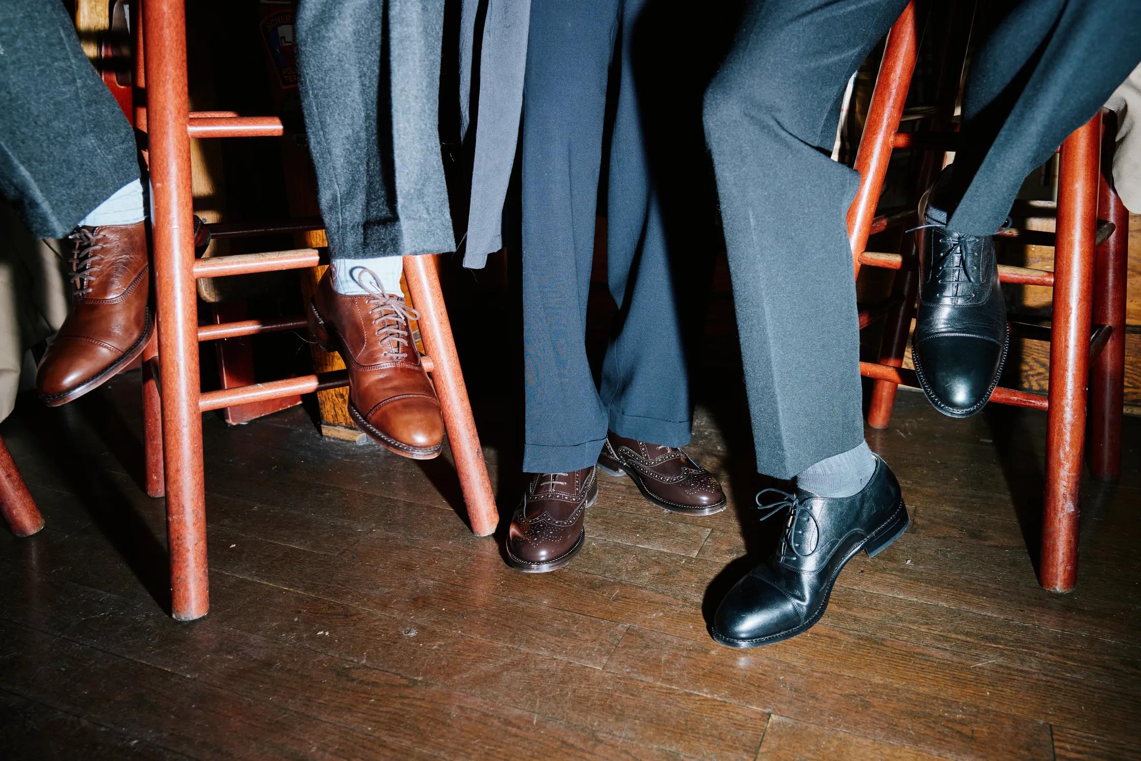 Three pairs of brown and black dress shoes under red stools on a wooden floor.