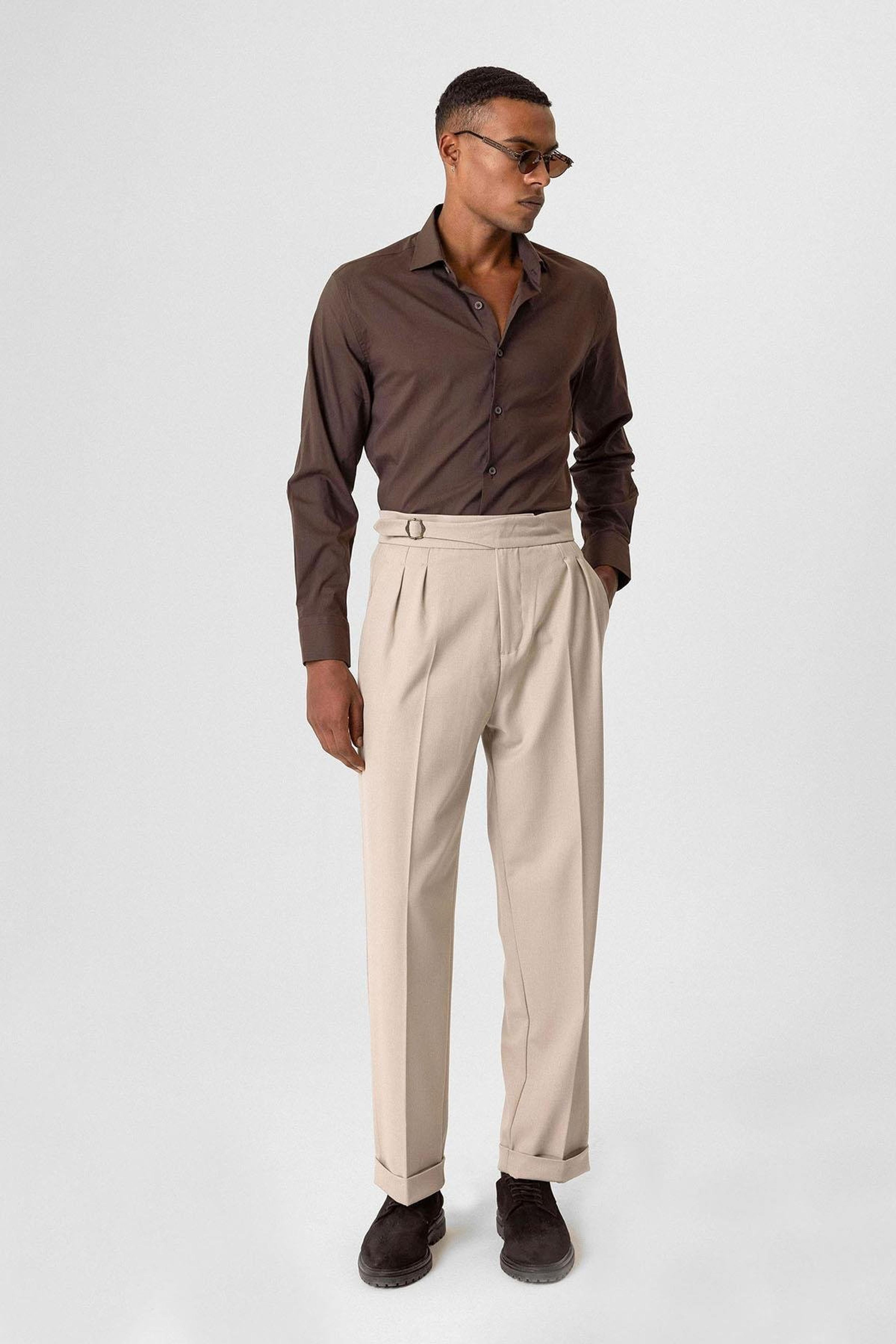 A man wearing a brown shirt, beige pleated trousers, and sunglasses stands against a light background.