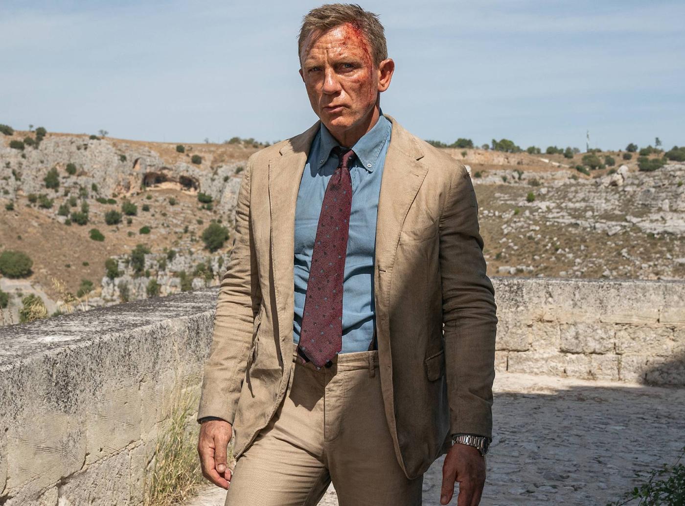 Daniel Craig with a bloody forehead, wearing a tan suit and tie, looking grim in a rocky landscape.