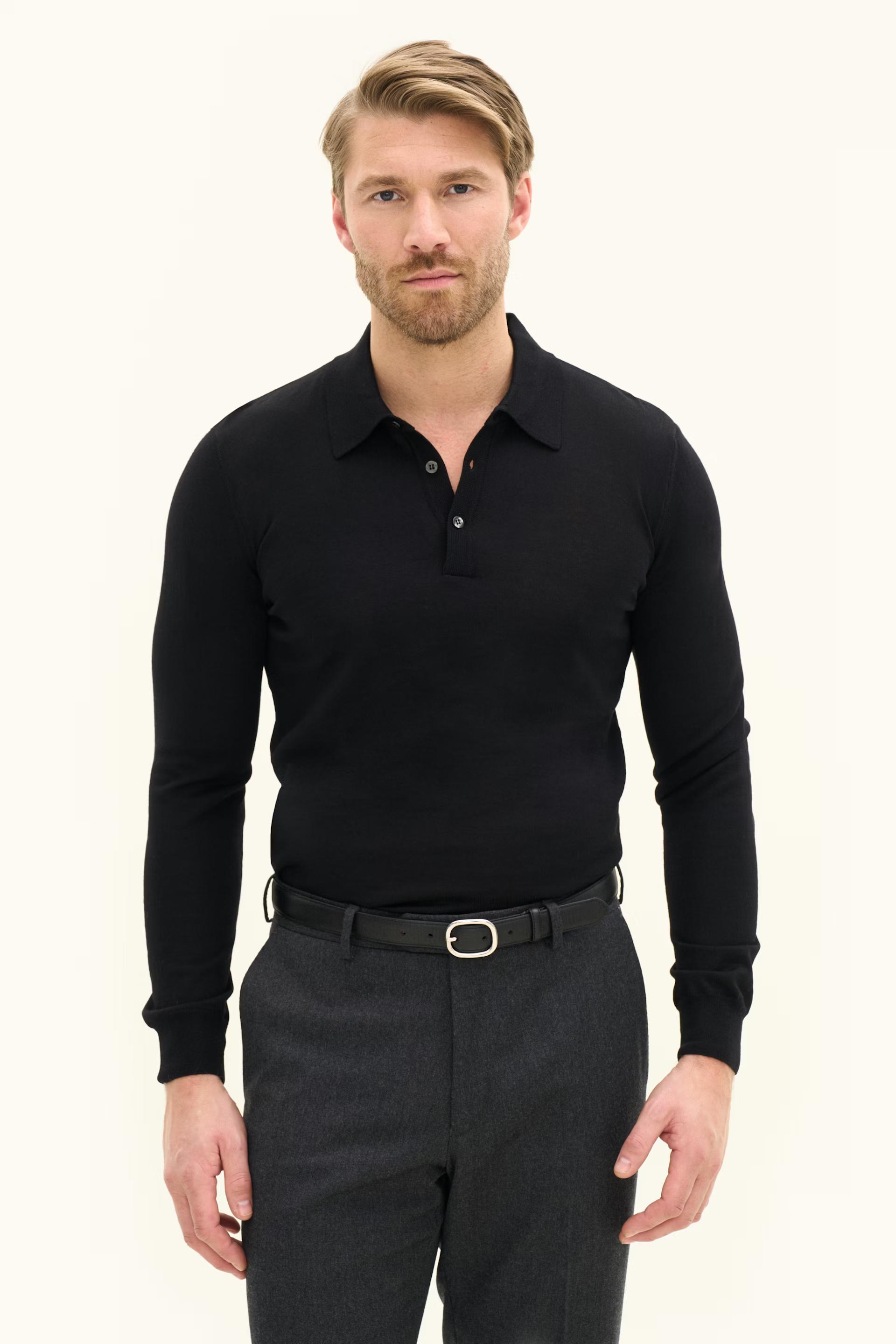 A man wearing a black long-sleeved knit polo shirt, dark gray trousers, and a black belt stands against a light background.