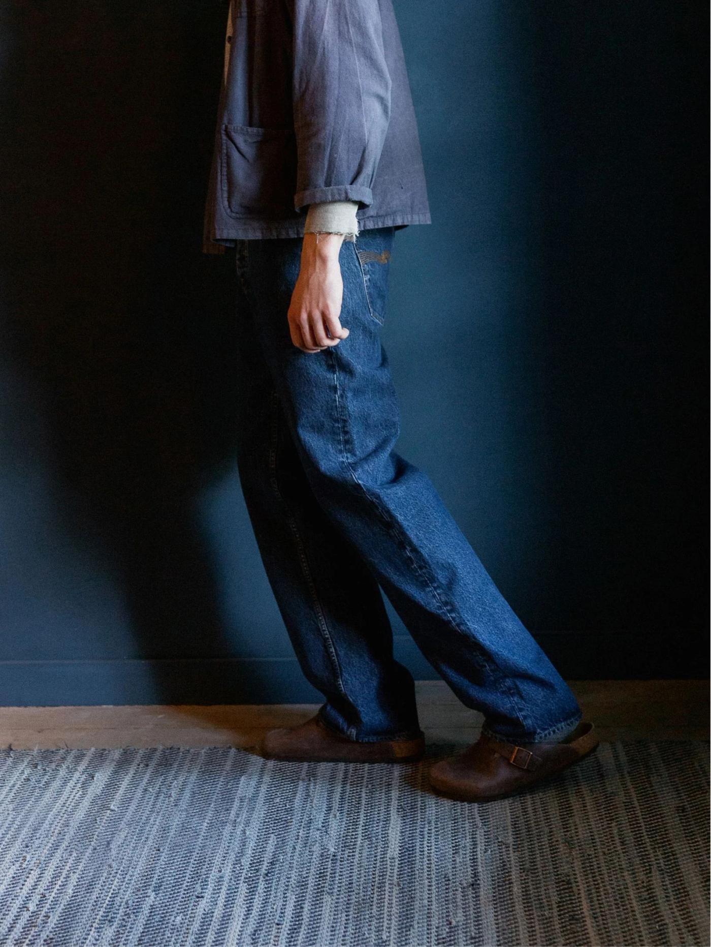 A person wearing a dark jacket, blue jeans, and brown clogs stands on a striped rug against a dark blue wall.