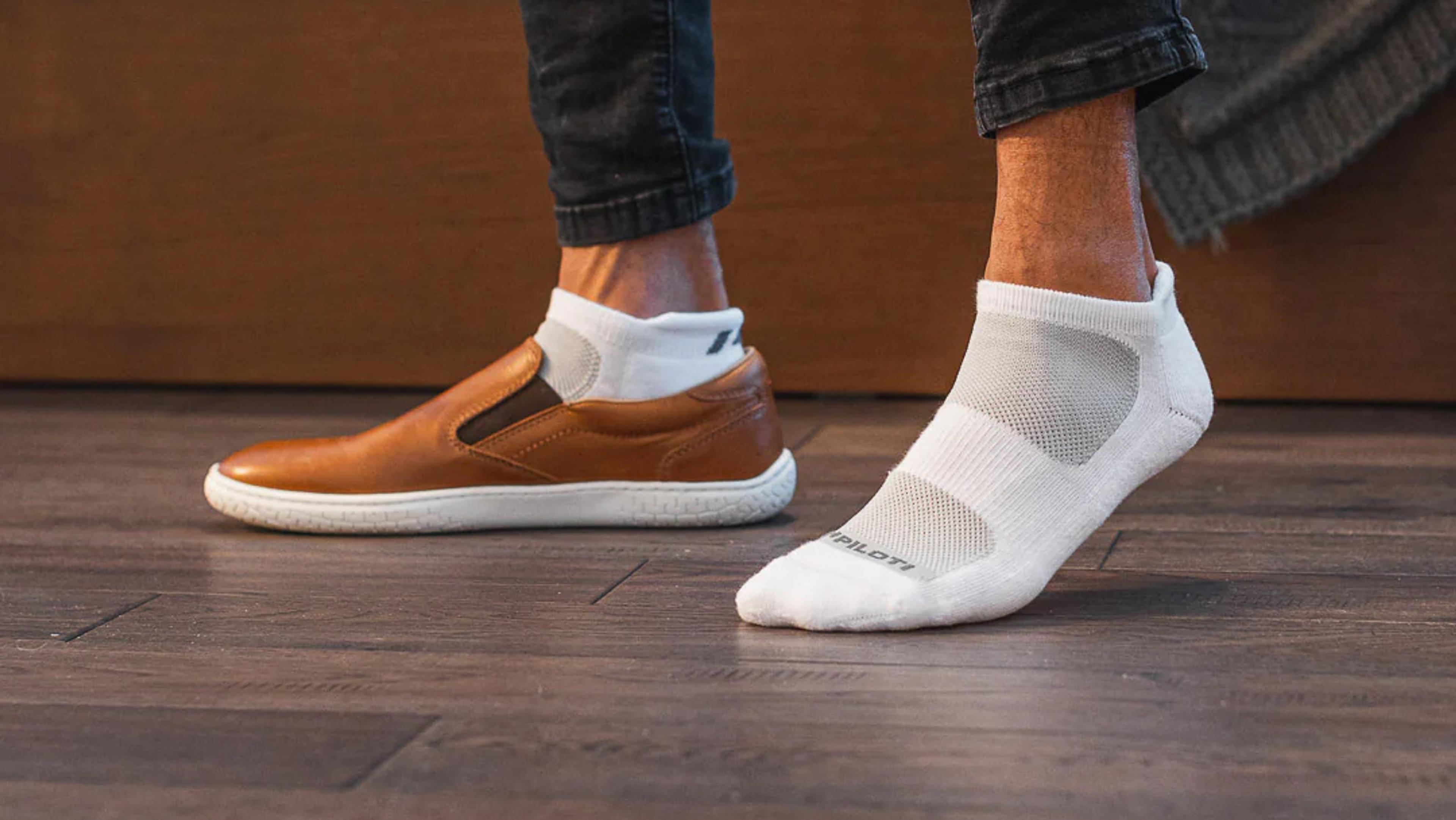 One foot in a brown slip-on shoe with a white sock, next to a foot wearing a white athletic sock on a wooden floor.