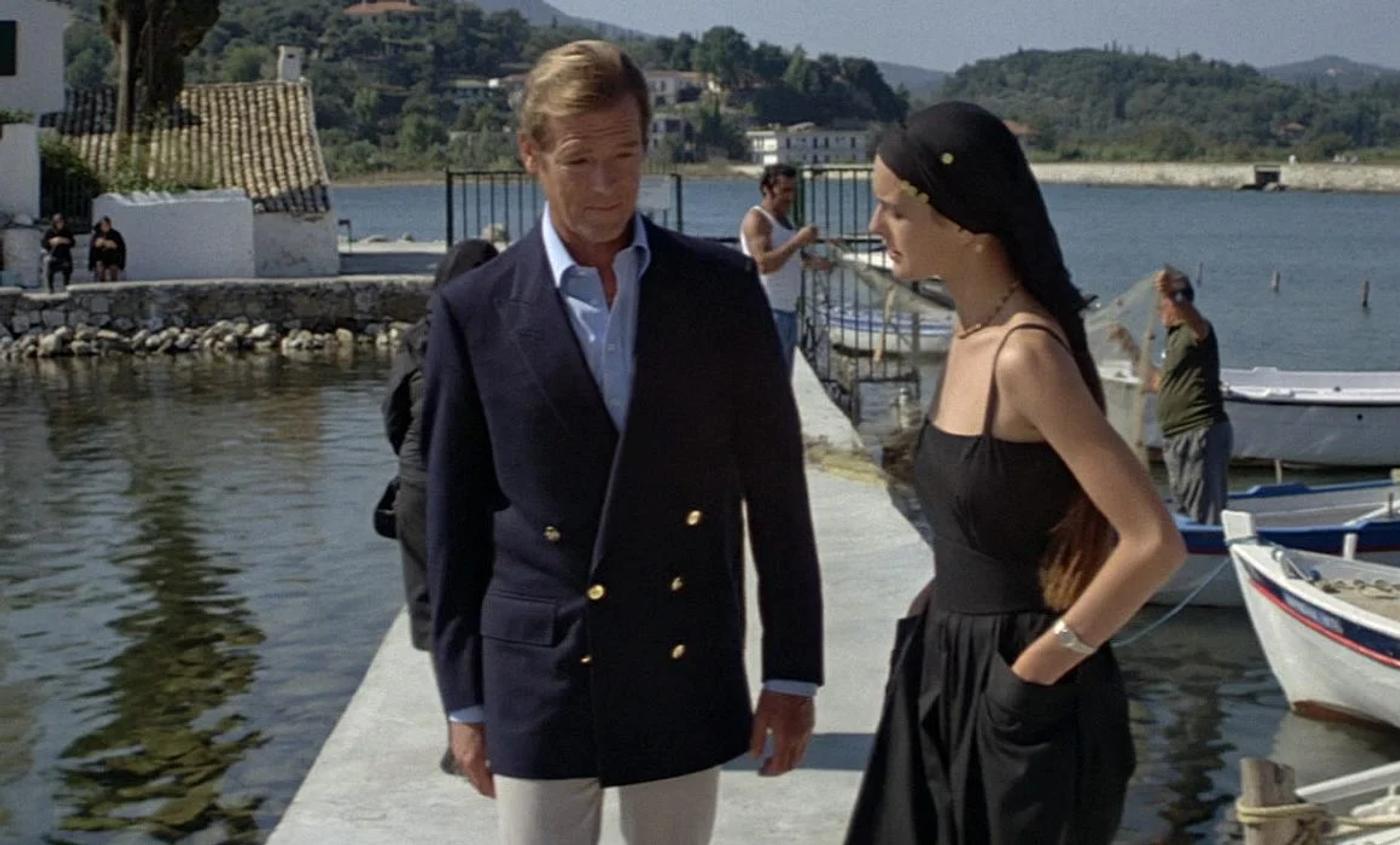 Roger Moore in a navy blazer and Carole Bouquet in a black dress and headscarf stand on a pier in a Mediterranean village.