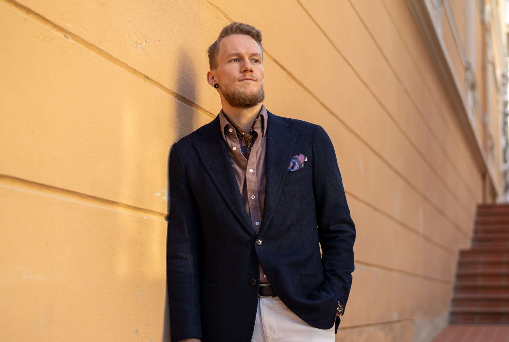 Man standing against wall wearing a blazer, shirt, neckerchief and white trousers.