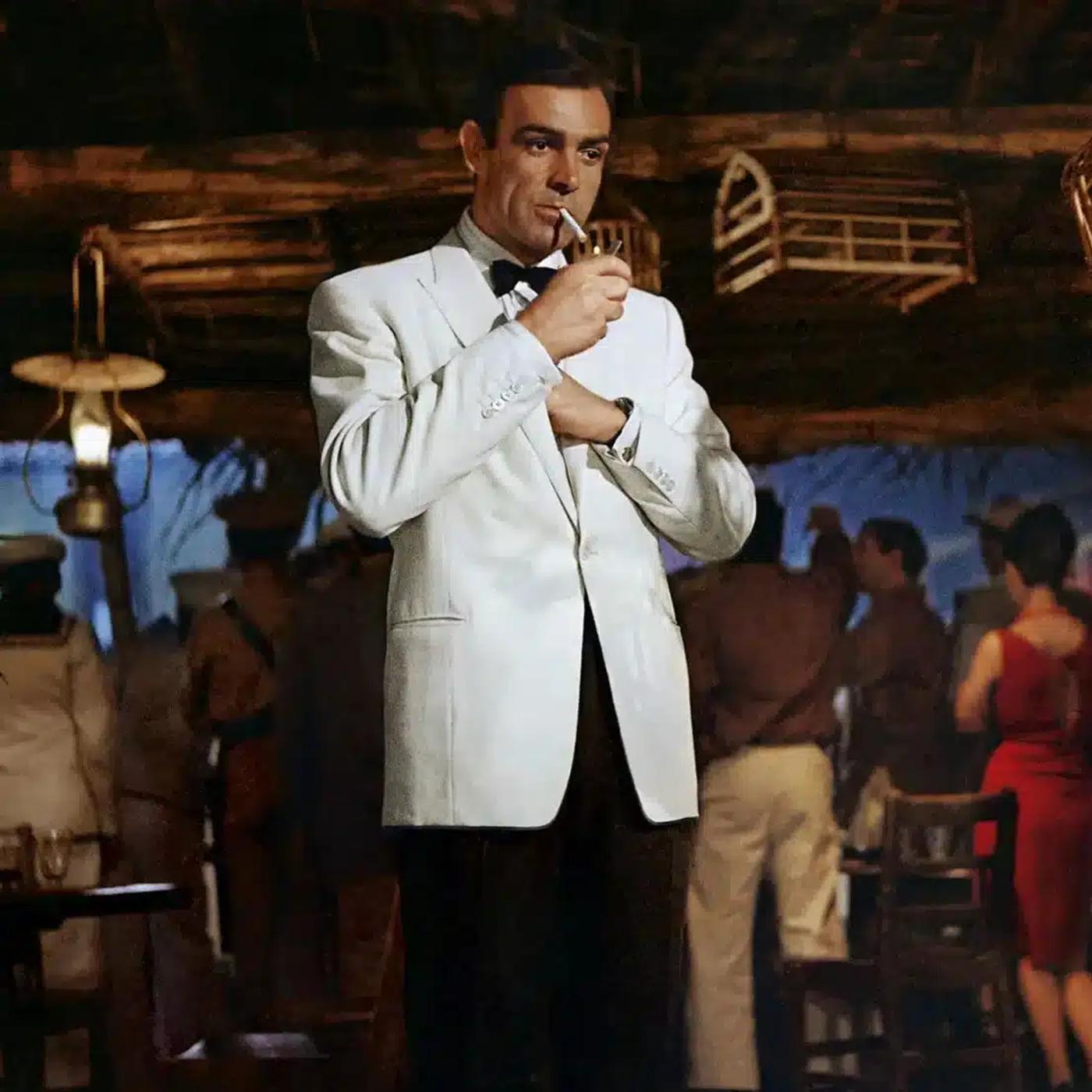 Sean Connery in a white dinner jacket lighting a cigarette in a crowded bar.