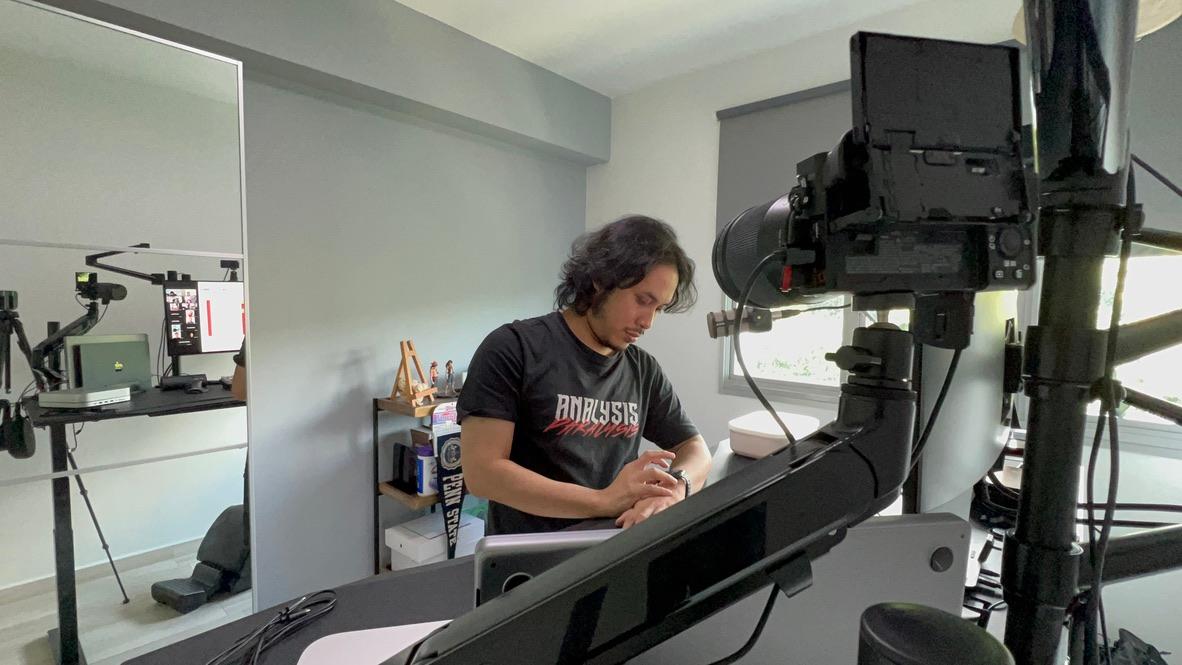 an wide-angle shot of ajmal standing up and looking at his watch while the there‘s a reflection of his setup on the mirror behind him