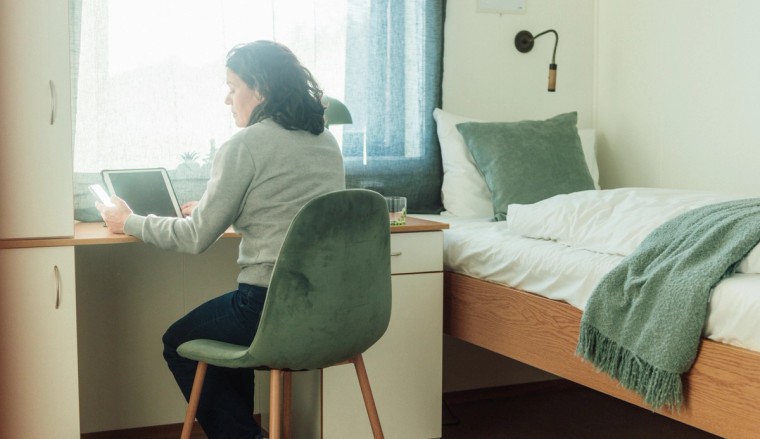 En kvinne sitter ved et skrivebord på et moderne rom og jobber på et nettbrett. Rommet har en seng, en stol og en lampe, med naturlig lys fra vinduet.