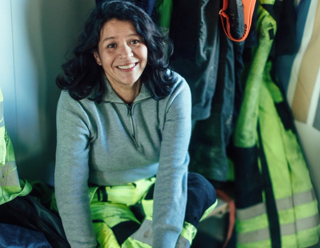 En smilende kvinne i arbeidsklær sitter i en garderobe, omgitt av refleksjakker og arbeidsutstyr.