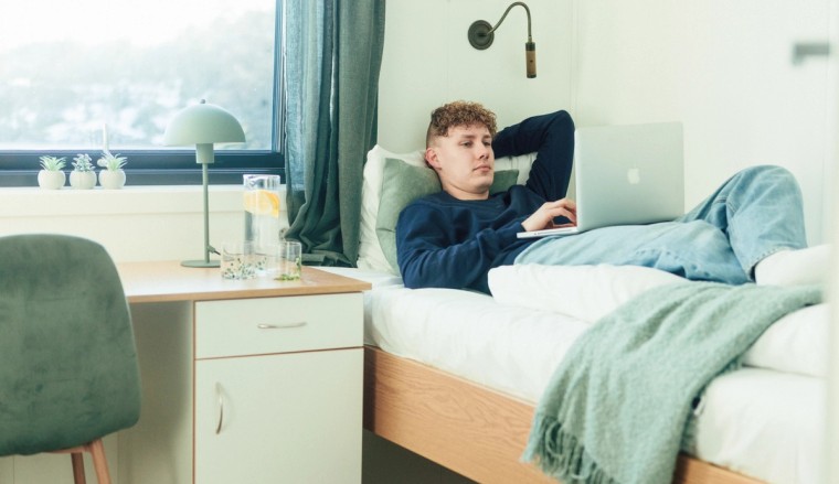 En ung mann ligger på en seng i et moderne, minimalistisk rom og jobber på en laptop. Rommet har et skrivebord, en stol, en nattlampe og et vindu med utsikt.