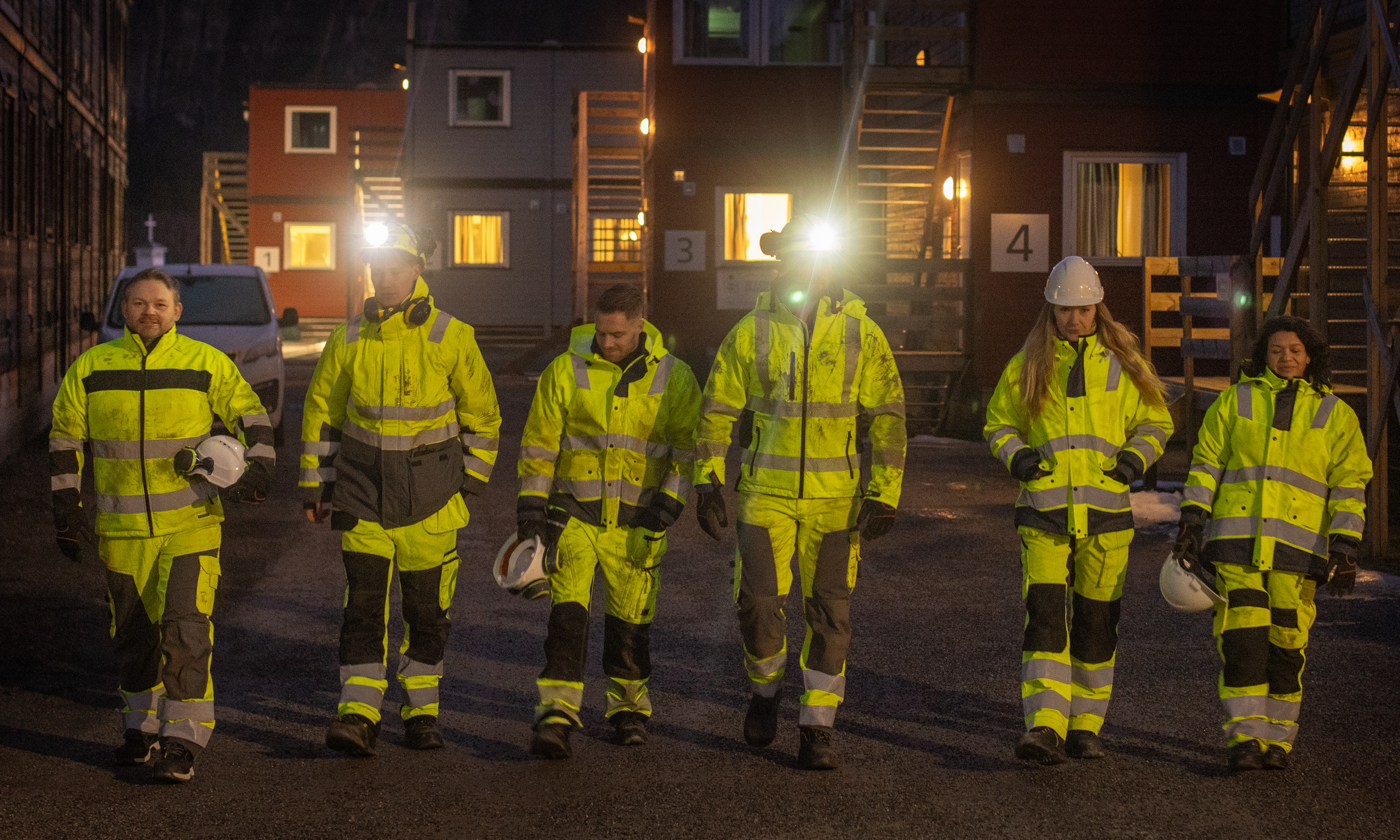 Fem anleggsarbeidere i synlige sikkerhetsklær går sammen på en arbeidsplass ved solnedgang, smilende og pratende. En lastebil og utstyr er i bakgrunnen.