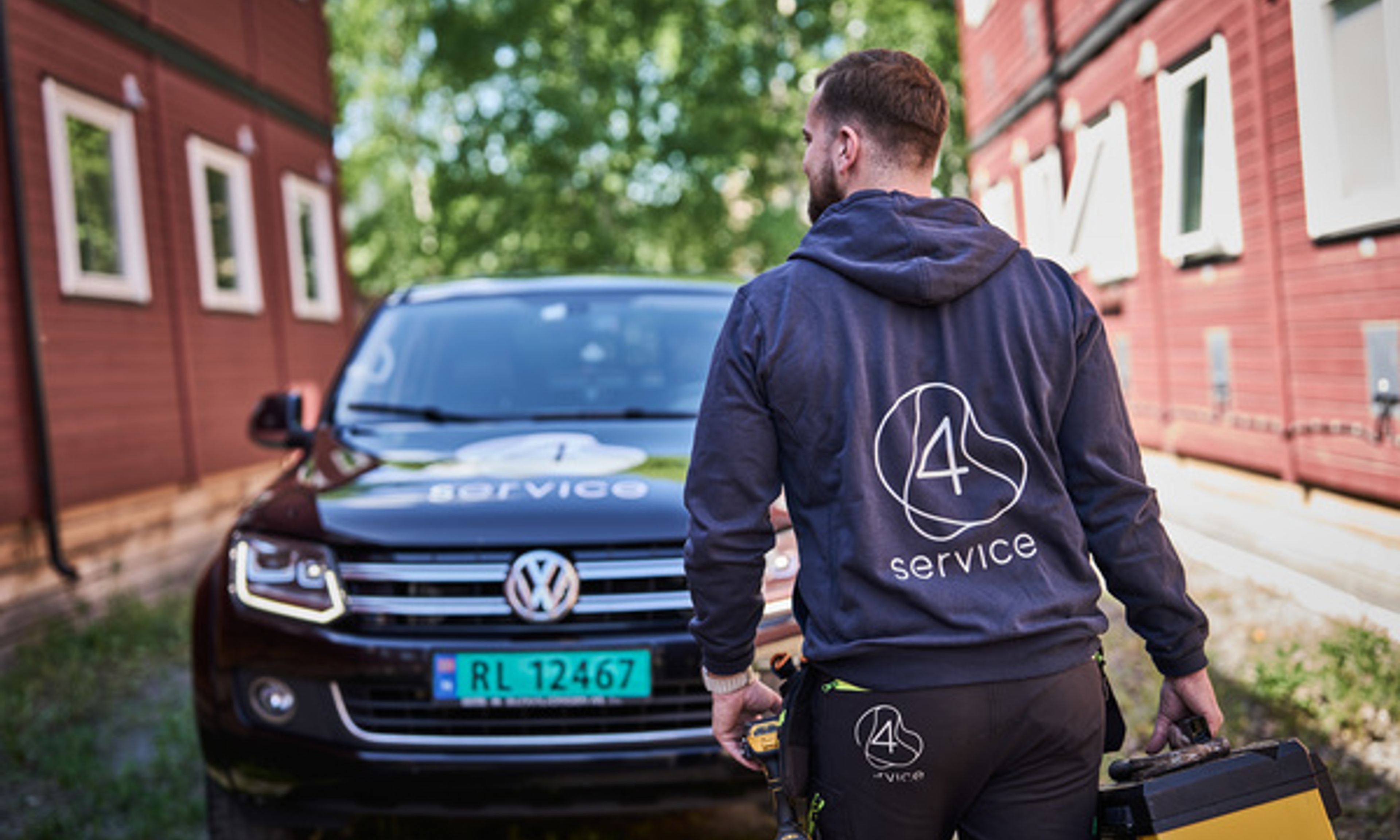En servicemedarbeider fra 4Service står med verktøykasse foran en svart Volkswagen med 4Service-logo, parkert mellom to røde bygninger på Camp Drammen.