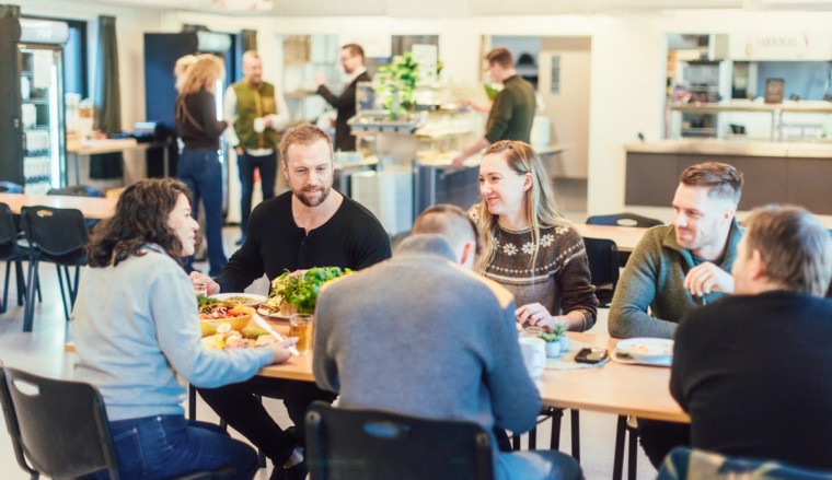 En gruppe mennesker nyter et måltid sammen i en lys og trivelig spisesal, mens andre gjester henter mat i bakgrunnen.