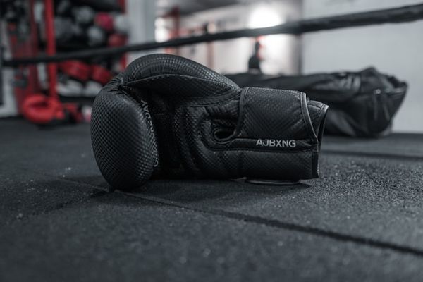 A black boxing glove with "AJBXNG" on the strap lies on a dark gym floor.