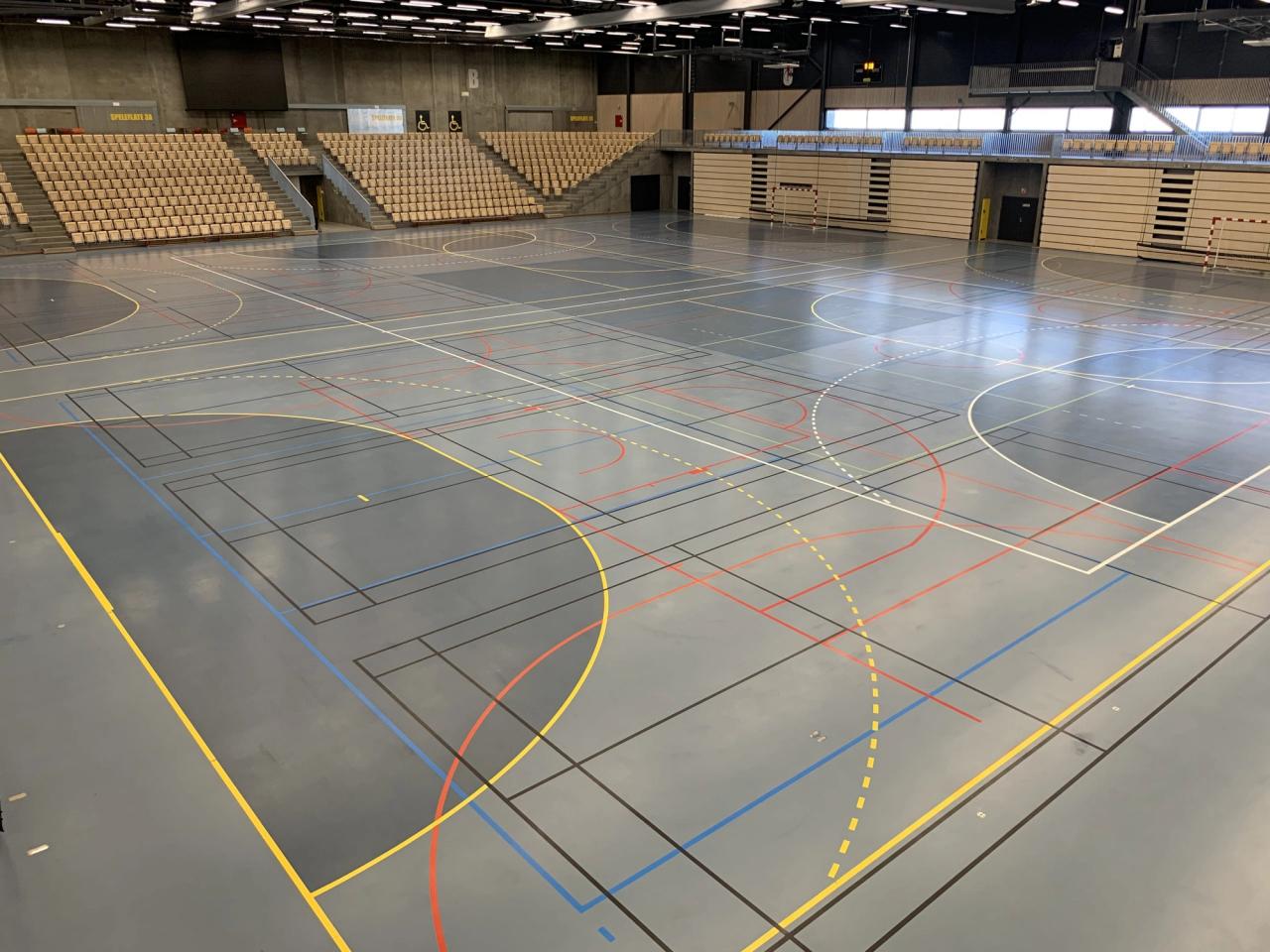 An empty indoor sports hall with a multi-sport court and tiered seating.