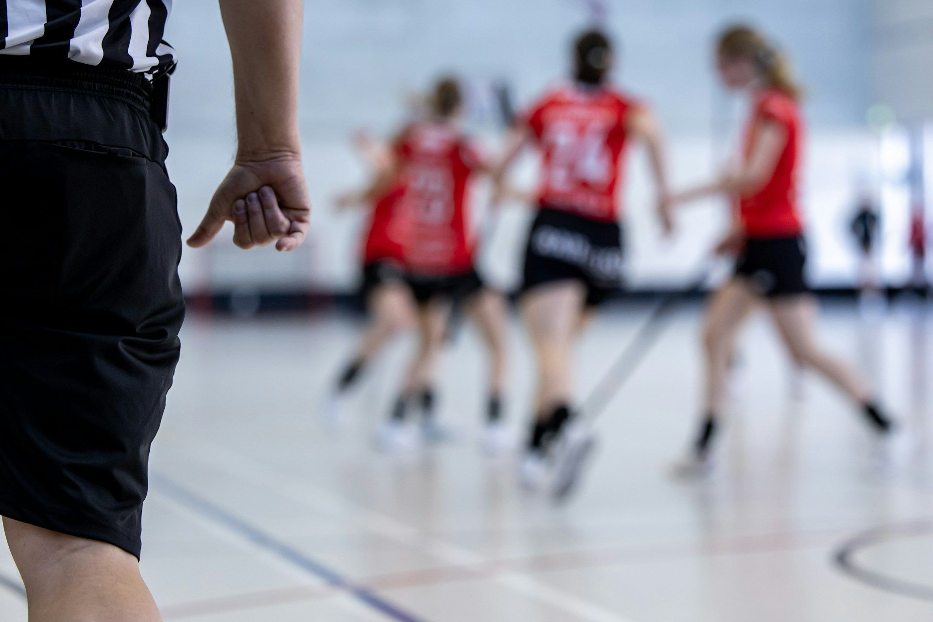 A referee from behind watches a blurred game of women's indoor stick sport.
