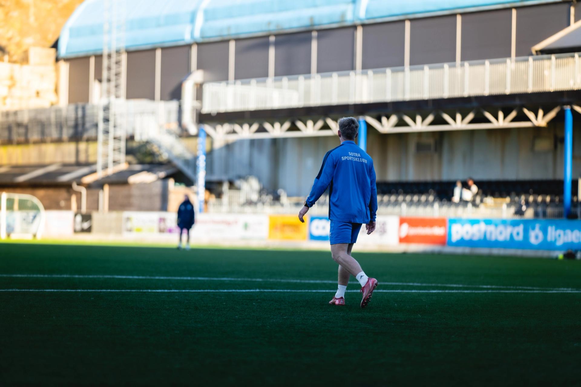 A person in a blue tracksuit walks on a green artificial turf field inside a stadium.