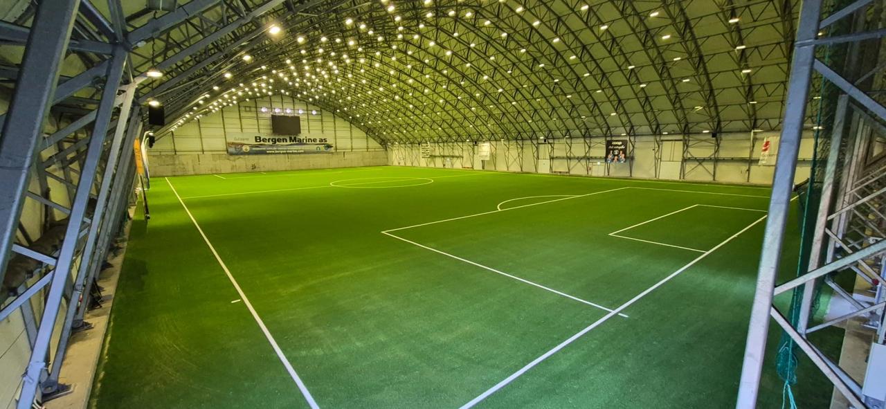 Empty indoor soccer field with green artificial turf under an arched, brightly lit ceiling.