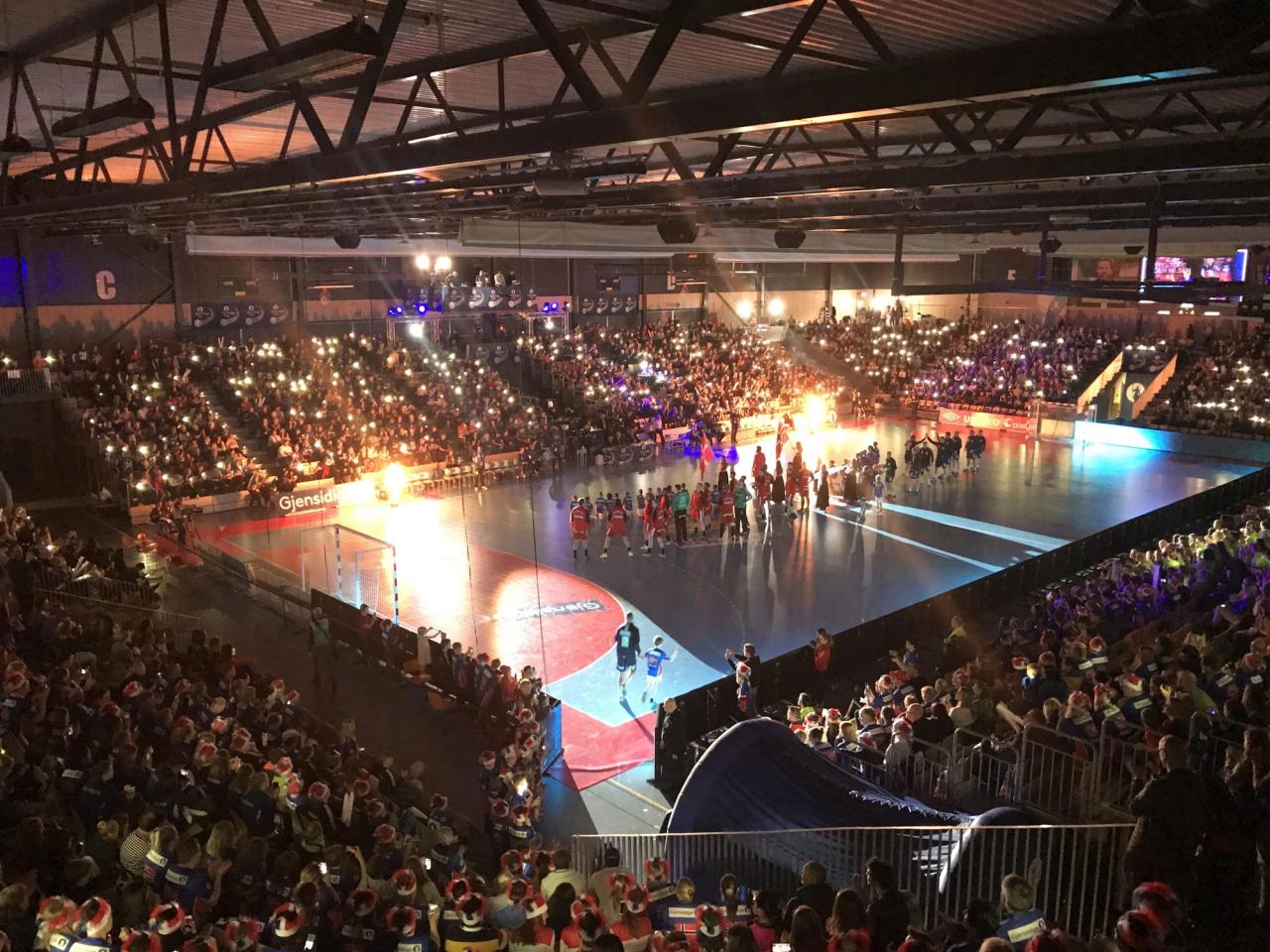 An indoor arena with a large crowd holding up lights, two teams on the court, and pyrotechnics.