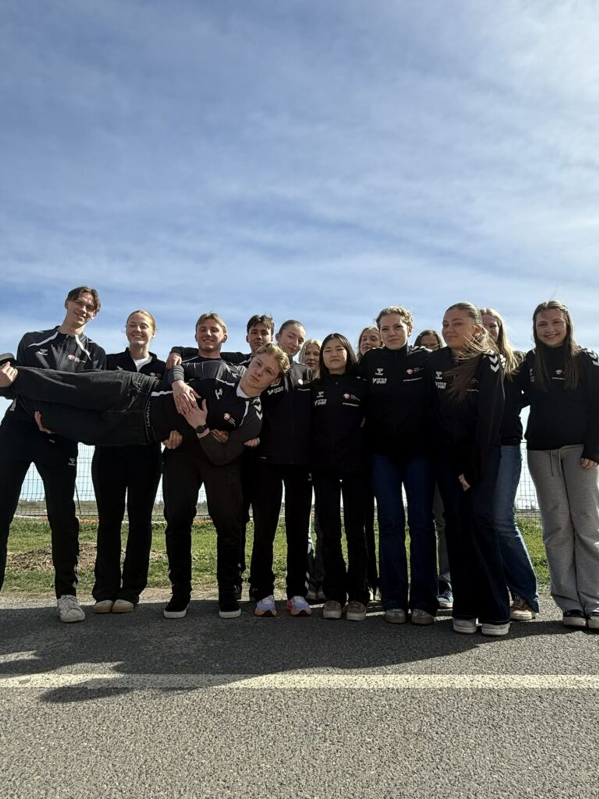 A group of smiling young people in athletic wear outdoors under a blue sky, with one person being carried horizontally.